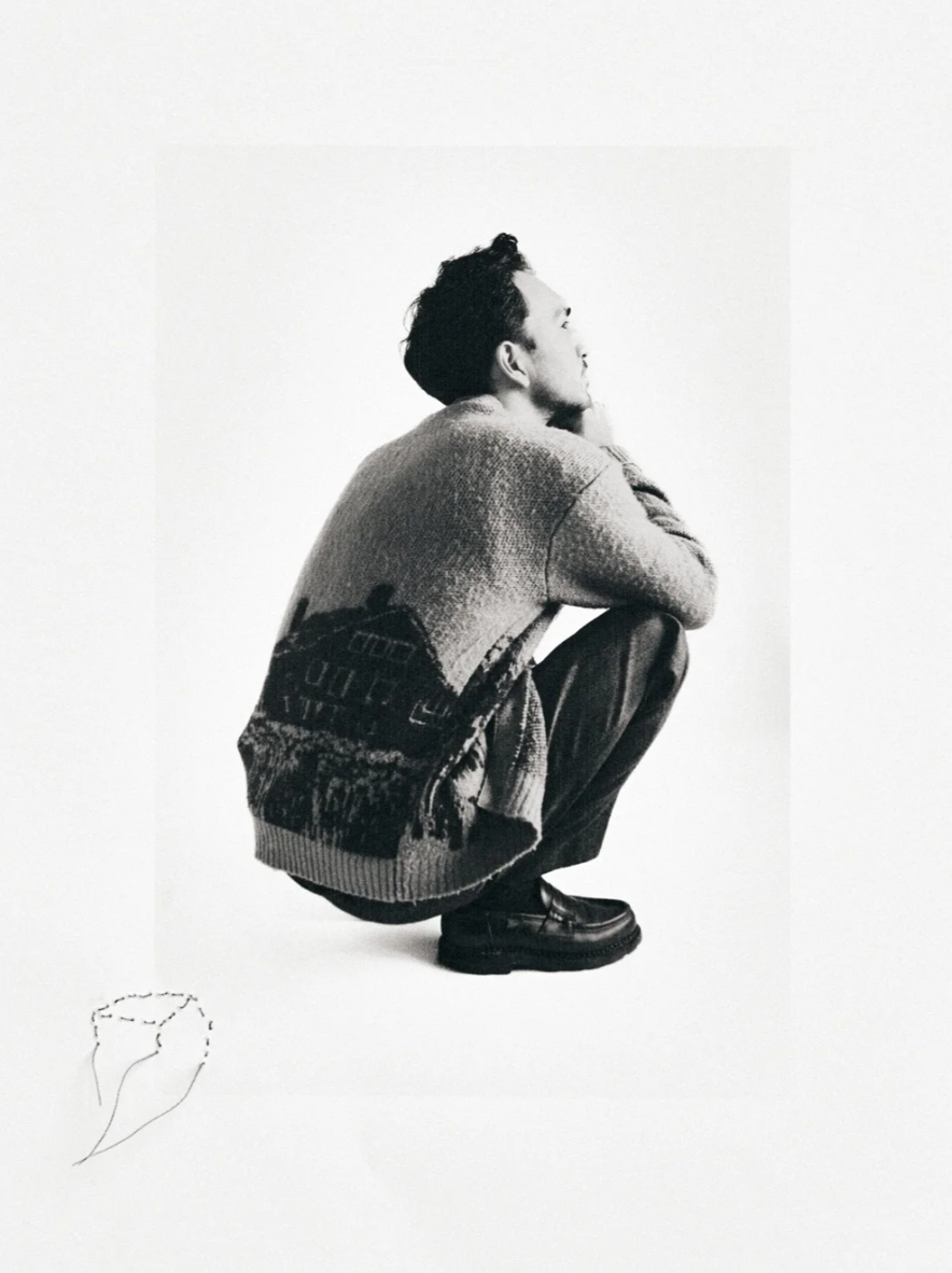 Black and white photo of a man squatting with his hand on his chin, wearing a sweater and dark pants, on a plain background. There is a sketch of a hand holding a partially cut gemstone or mineral in the bottom left corner.