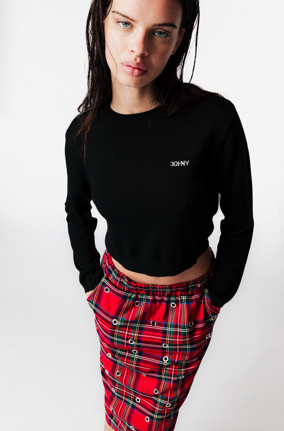Young woman with dark hair and blue eyes wearing a black crop top with white text and red plaid skirt with eyelet details, standing against a plain white background.