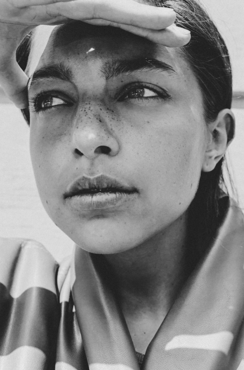 A black and white close-up photo of a young woman with freckles, looking to the side, holding her hand above her forehead, with a serious expression on her face.