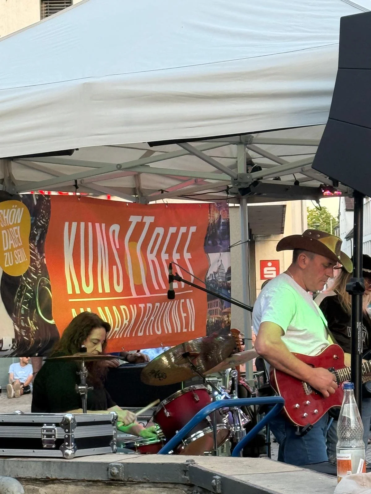 Musiker spielt Gitarre vor einem orangefarbenen Banner bei einer outdoor Veranstaltung, weitere Musiker im Hintergrund, auf einem Stadtplatz in Deutschland.