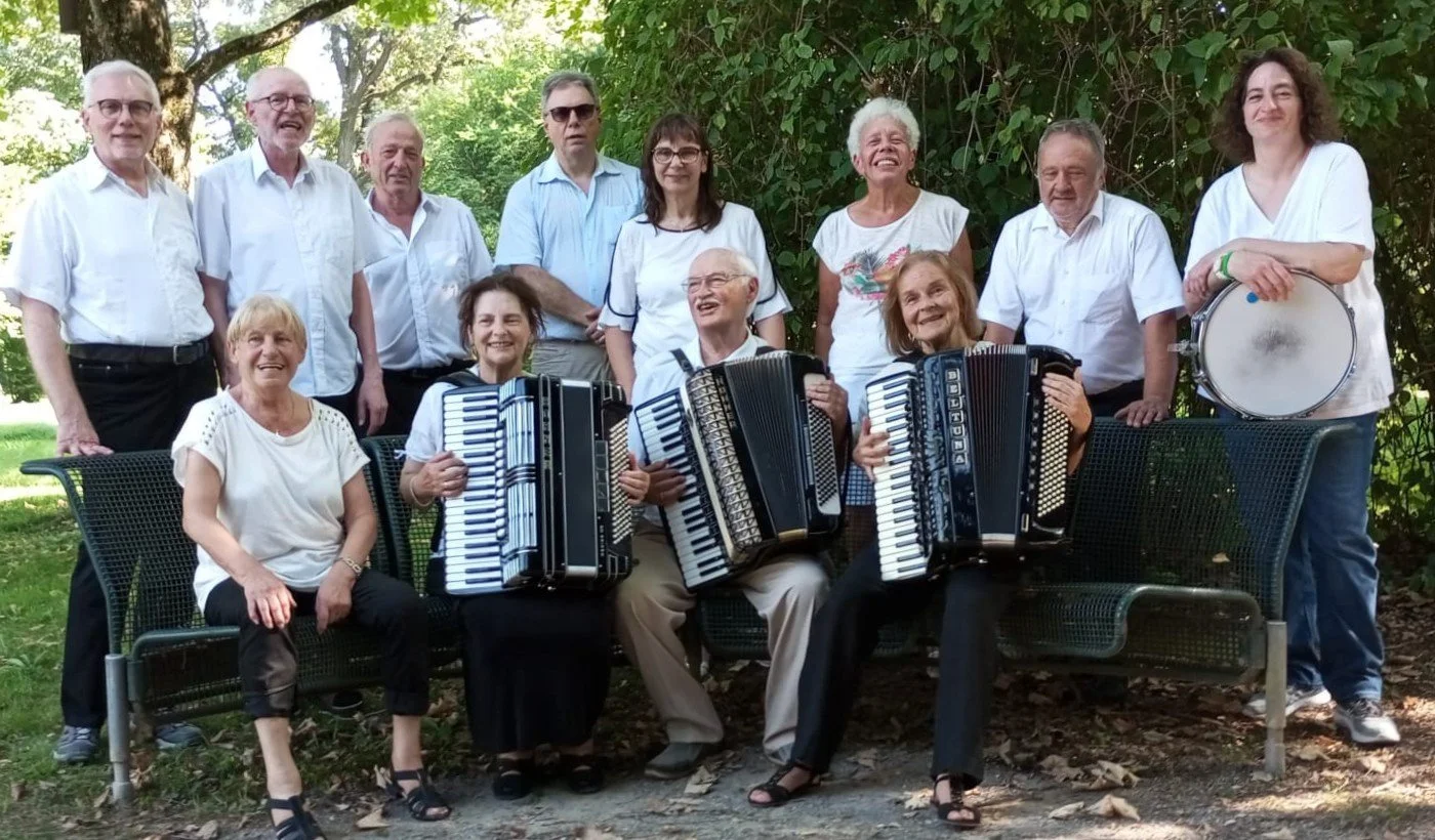 Eine Gruppe von Erwachsenen mit Musikinstrumenten, darunter Akkordeons und eine Trommel, posiert im Freien vor Bäumen und grünem Laub.