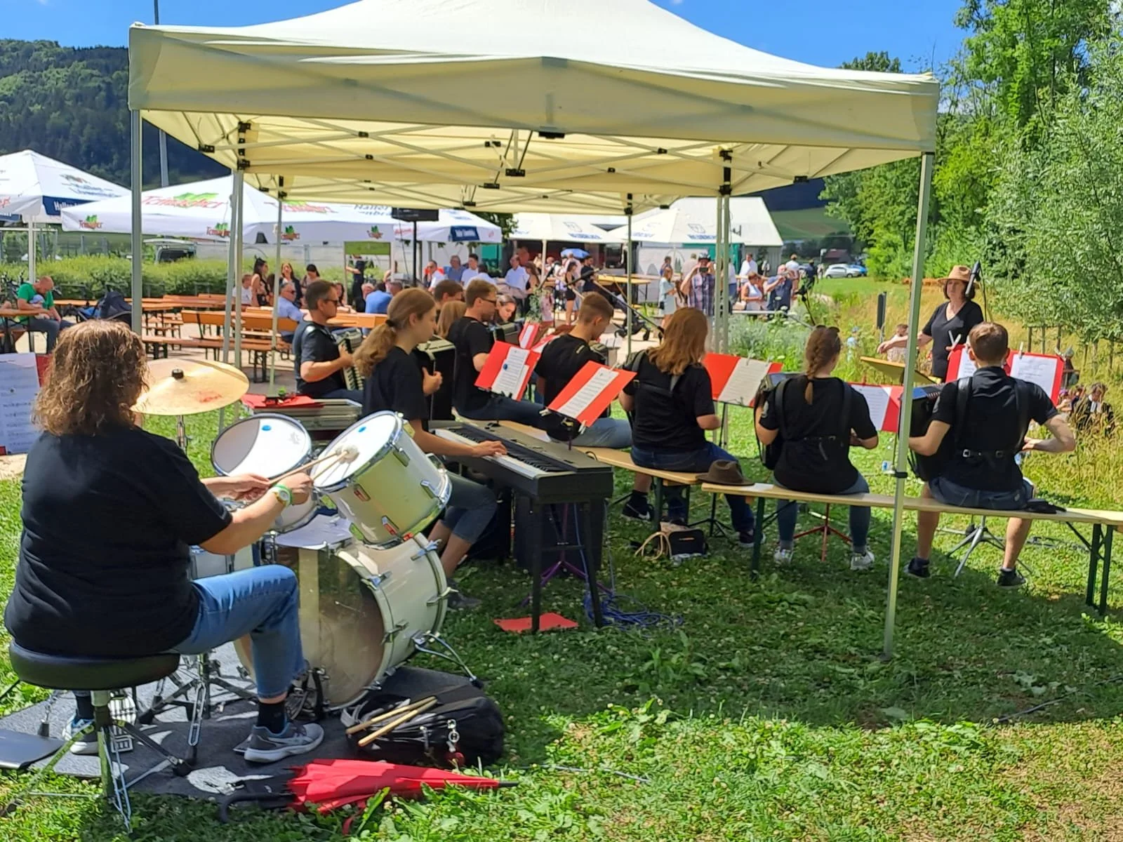 Eine Band spielt im Freien unter einem Zelt bei sonnigem Wetter, umgeben von grünen Bäumen und einem Himmel mit wenigen Wolken.