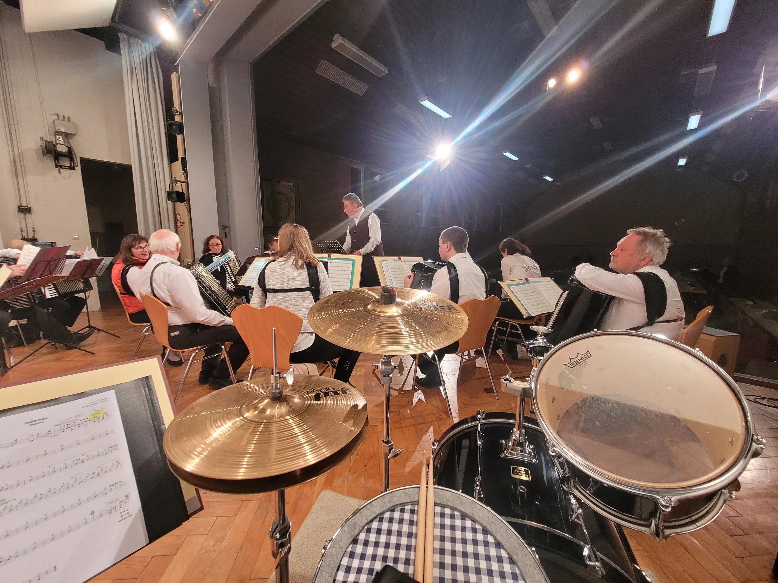 Eine Bühne mit Musikern, die sich auf eine Aufführung vorbereiten. Im Vordergrund sind Schlaginstrumente wie Trommeln sichtbar, im Hintergrund sitzen Musiker mit Notenblättern. Ein Dirigent steht in der Mitte, beleuchtet von Scheinwerfern.