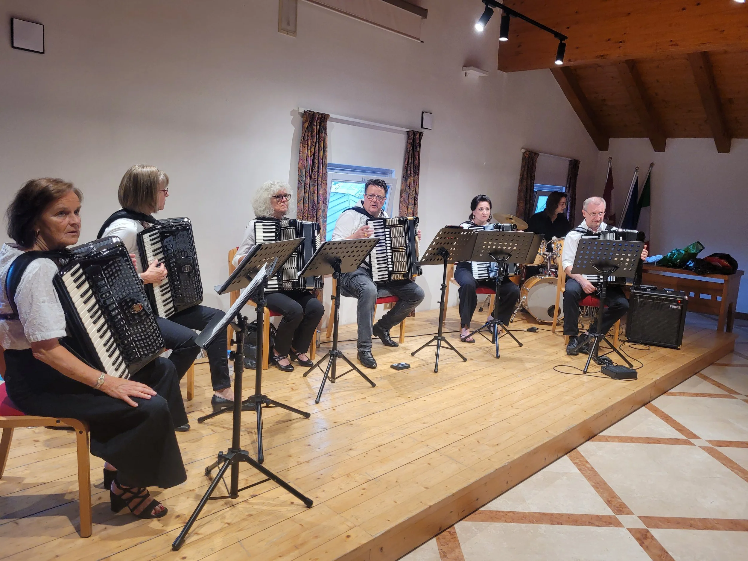 Gruppe von Musikern, die Akkordeons, Trommeln und andere Instrumente auf einer Bühne in einem Saal spielen, mit Vorhängen im Hintergrund.