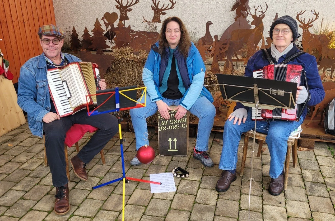 Drei Musiker sitzen auf Stühlen auf einer gepflasterten Terrasse, mit einem Hintergrund aus Metallfiguren von Rehen und Tieren. Zwei der Musiker spielen Akkordeon, die Frau in der Mitte sitzt und lächelt. Musikständer, eine Notenliste, eine rote Weihnachtsbaumkugel, ein Halfter mit kleinen Glocken und eine Tafel sind sichtbar.