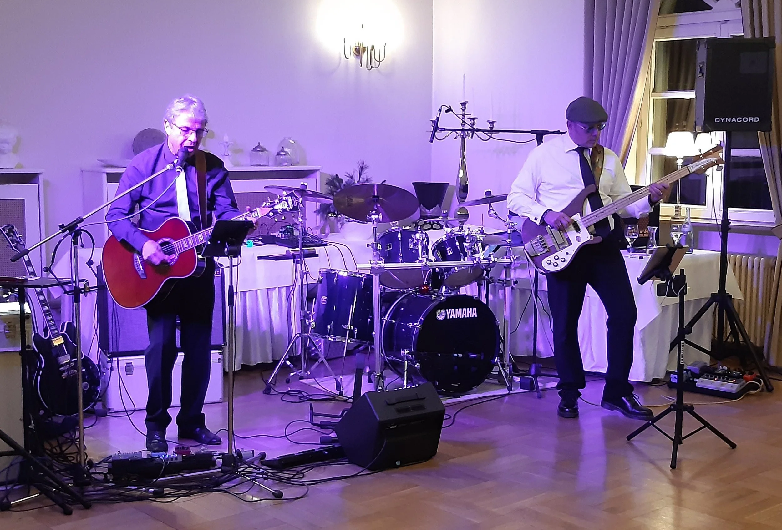 Zwei Musiker spielen in einem Innenraum: einer mit einer Gitarre, der andere mit einem Bass. Es gibt ein Schlagzeug und Verstärker im Hintergrund, sowie Kerzen und Dekorationen auf den Möbeln. Die Szene ist schwach beleuchtet mit blauem Licht.
