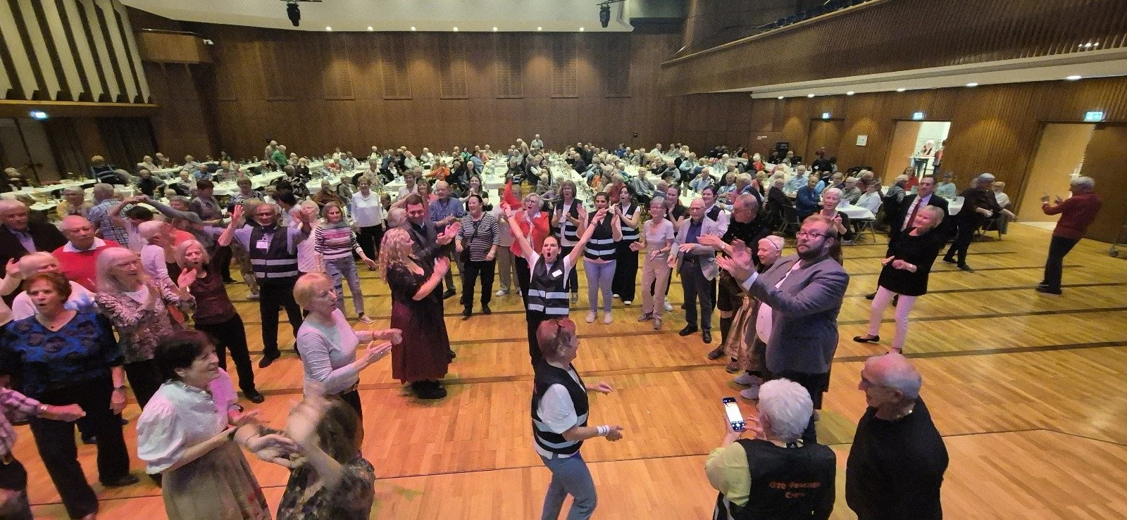 Menschen tanzen und feiern in einer großen Halle mit Holzfußboden und dunklen Wänden.
