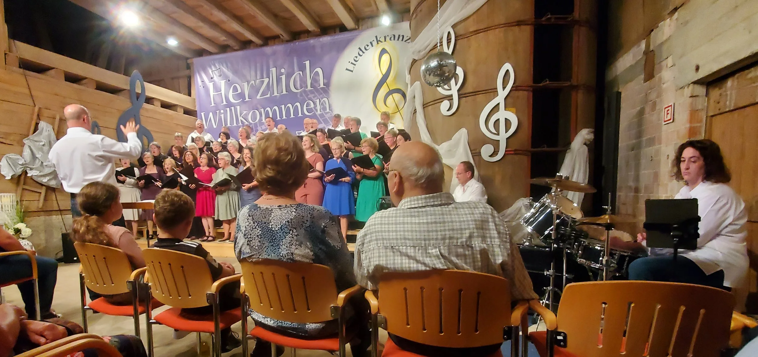 Ein Chor singt auf einer Bühne während einer Aufführung. Ein Dirigent steht vor dem Chor, der aus Männern und Frauen besteht. Im Hintergrund ist eine große silberne musikalische Note und ein Banner mit dem Text "Herzlich Willkommen" und "Liederkranz" zu sehen. Es sitzen mehrere Zuschauende vor der Bühne, während neben dem Chor Musiker mit Schlagzeugen sitzen.