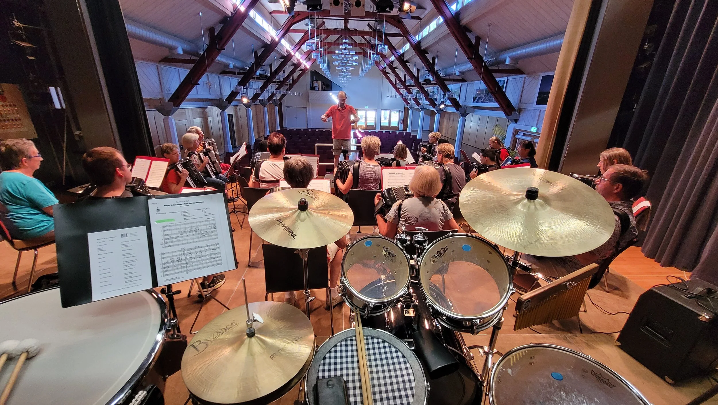 Auftritt, bei der ein Dirigent am Dirigieren ist. Musiker sitzen mit ihren Instrumenten, darunter auch Akkordeons. Im Vordergrund sind Schlagzeuge zu sehen. Der Raum hat eine hohe Decke mit Holzbalken und große Fenster, die für viel Licht sorgen.