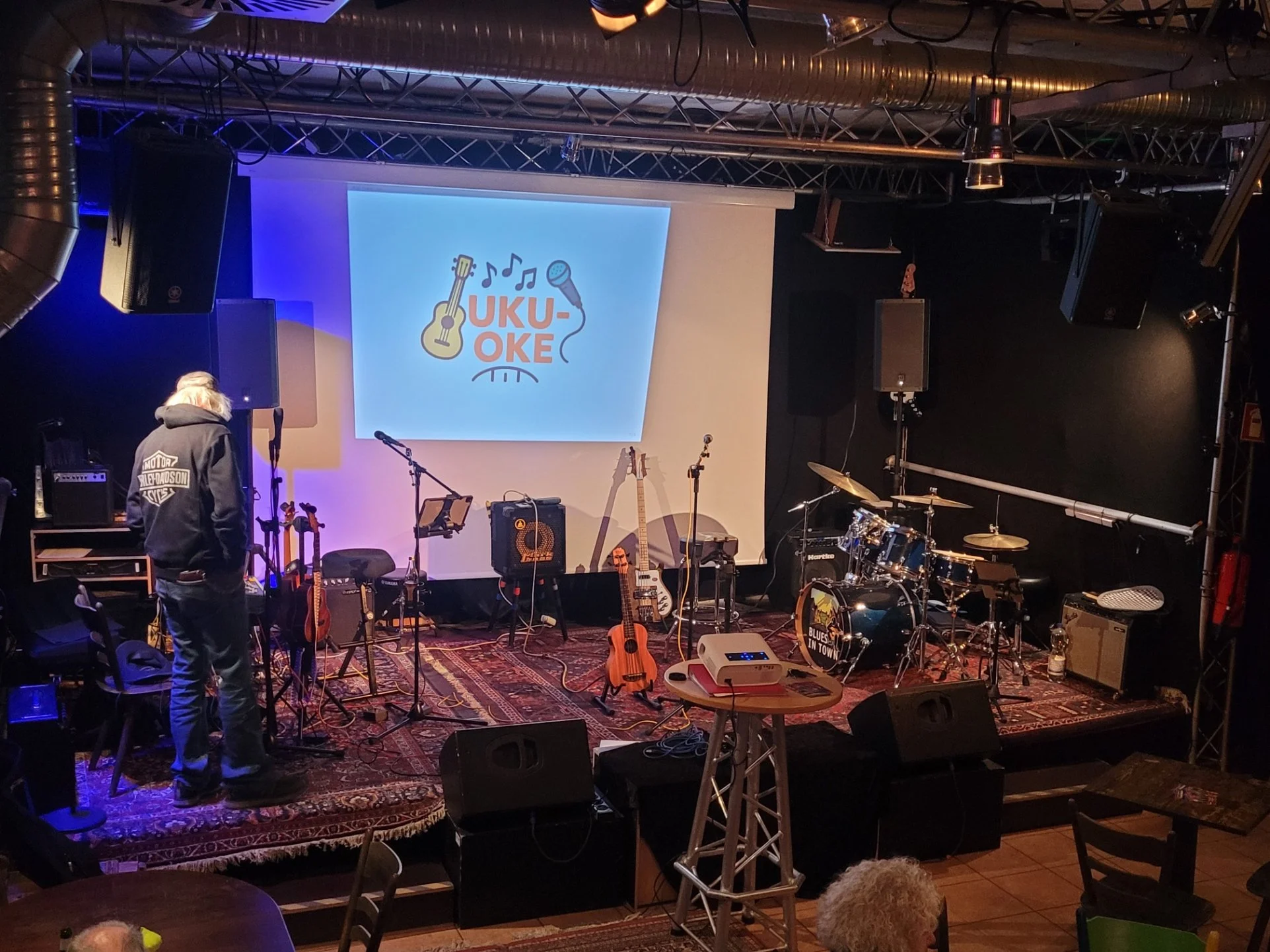 Bühne mit Musik- und Instrumentszubehör, darunter Gitarren, Mikrofone, Verstärker und Schlagzeug, mit einem großen Bildschirm im Hintergrund, auf dem das Logo "UKU-OKE" zu sehen ist, bei einer Veranstaltung in einem Musikclub