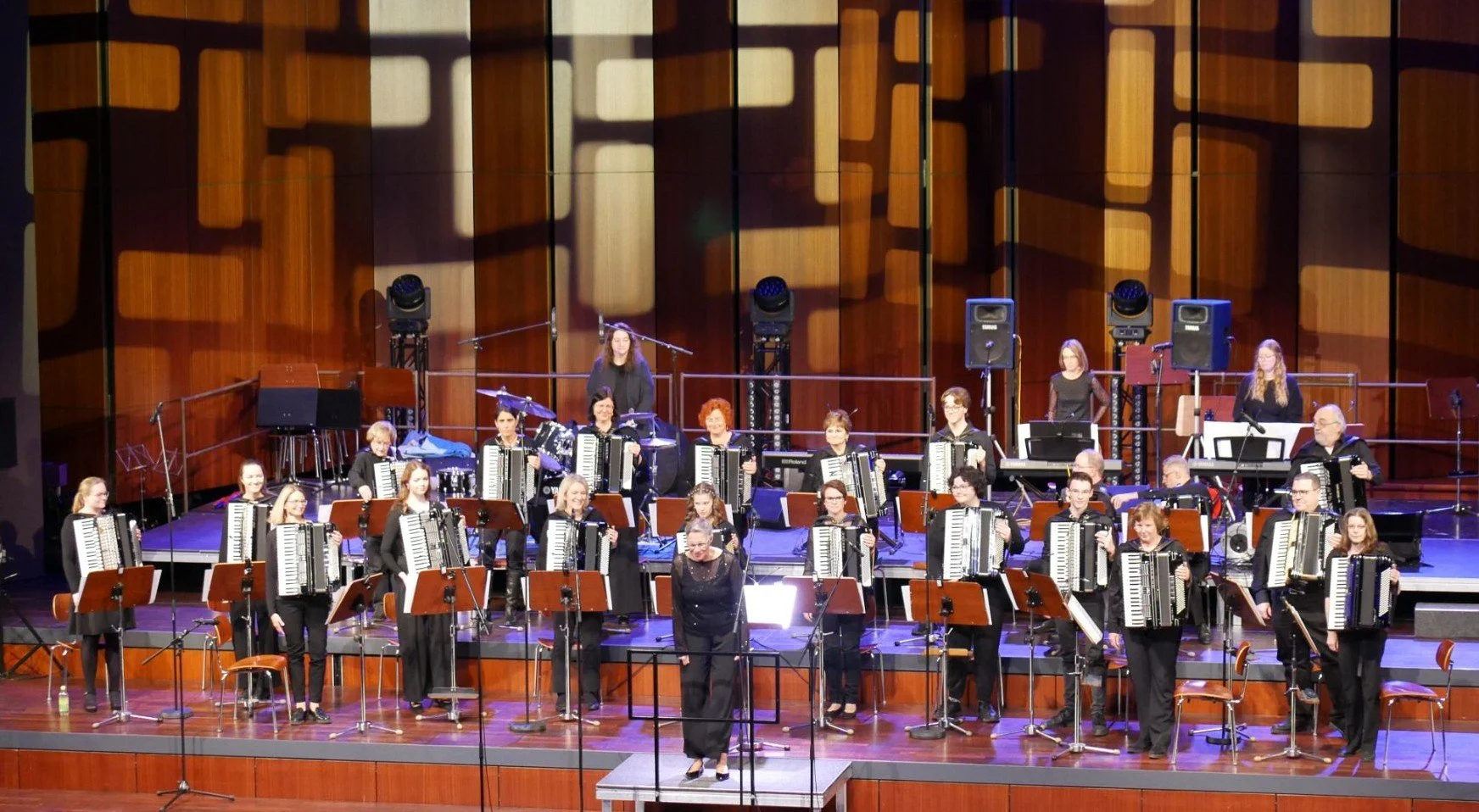 Ein Orchester auf einer Bühne, bestehend aus mehreren Musikerinnen und Musikern, die Akkordeons spielen, mit einem Dirigenten vorne. Im Hintergrund befinden sich Lautsprecher und Lichttechnik, und die Bühne ist mit Holzpaneelen verkleidet.