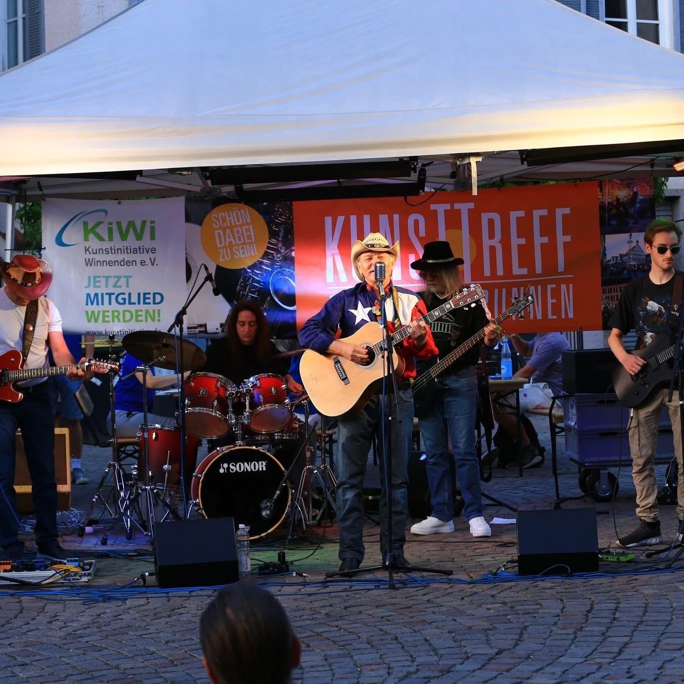 Eine Band spielt auf einer Bühne unter einem weißen Zelt. Es sind mehrere Musiker mit Gitarren, Schlagzeug und Mikrofon zu sehen, im Hintergrund ein rotes Banner mit dem Text "Kunsttreff".