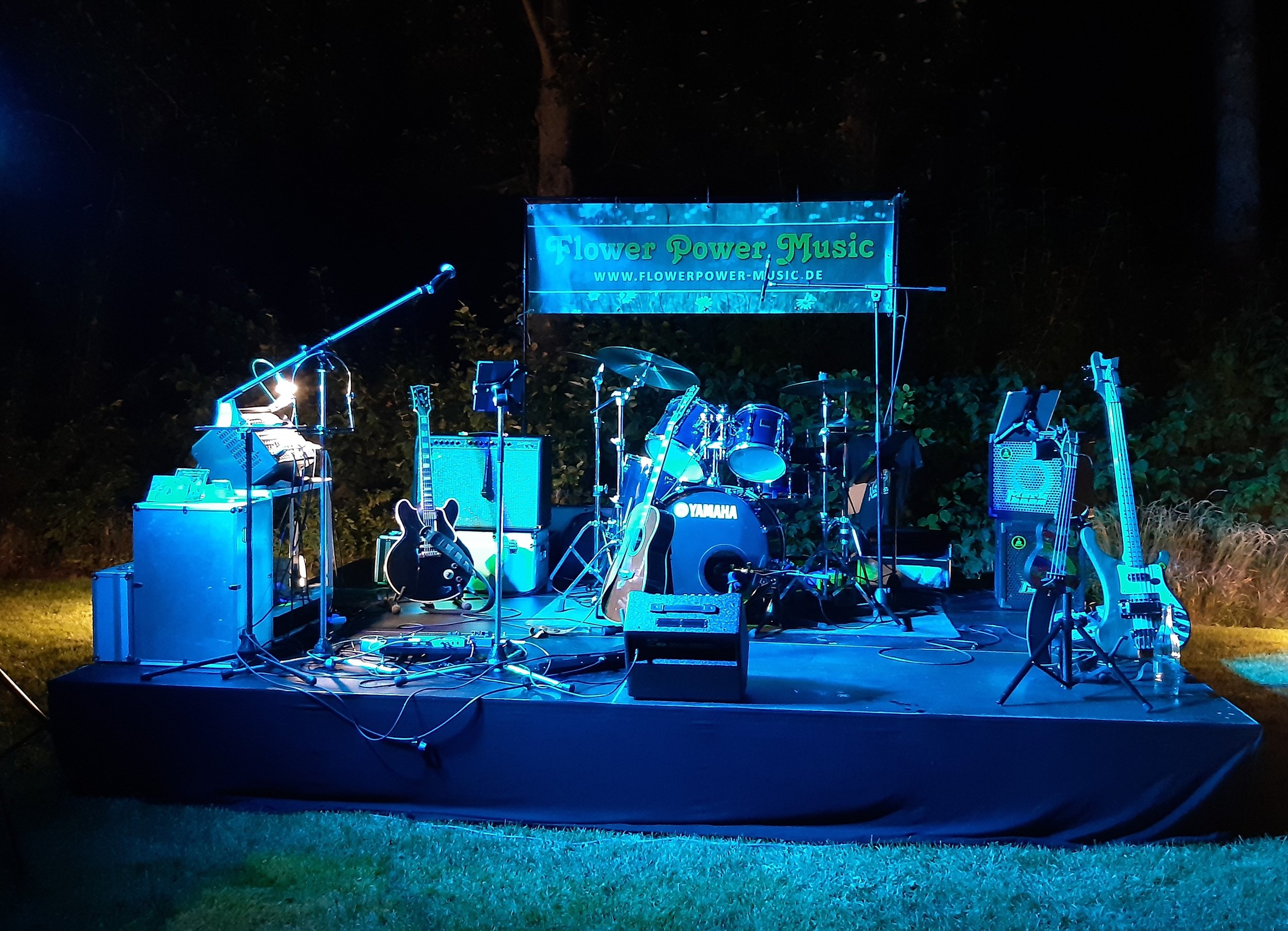 Eine leere Musikbühne im Freien bei Nacht mit Instrumenten und Ausrüstung, Beleuchtung in Blau, ein Banner mit der Aufschrift 'Flower Power Music', umgeben von Bäumen.