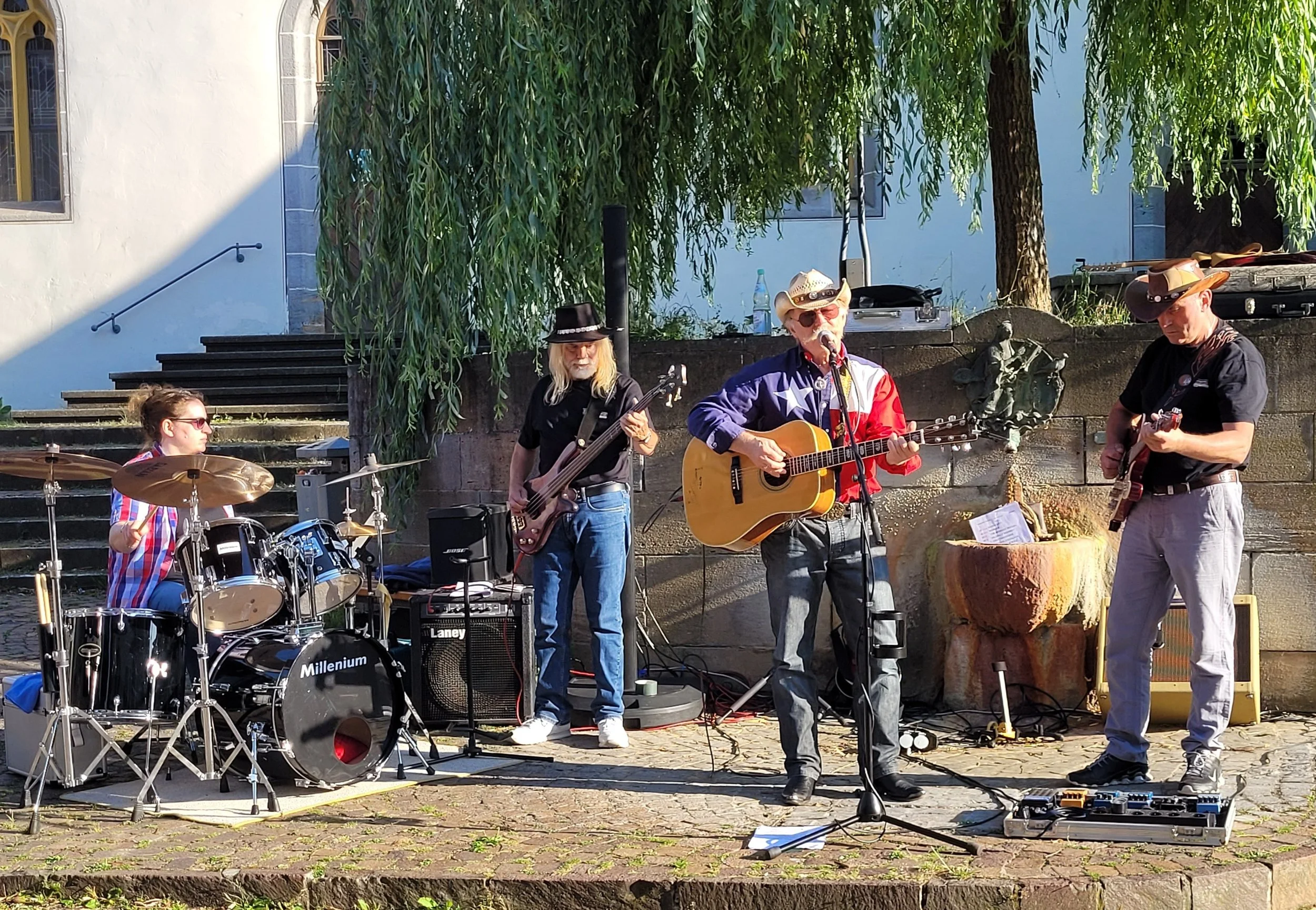 Eine Gruppe von Musikern, darunter ein Schlagzeuger, ein Bassgitarrist, ein Sänger mit Gitarre und ein weiterer Gitarrist, spielt auf einer kleinen Bühne im Freien, umgeben von Bäumen und einem weißen Gebäude im Hintergrund.