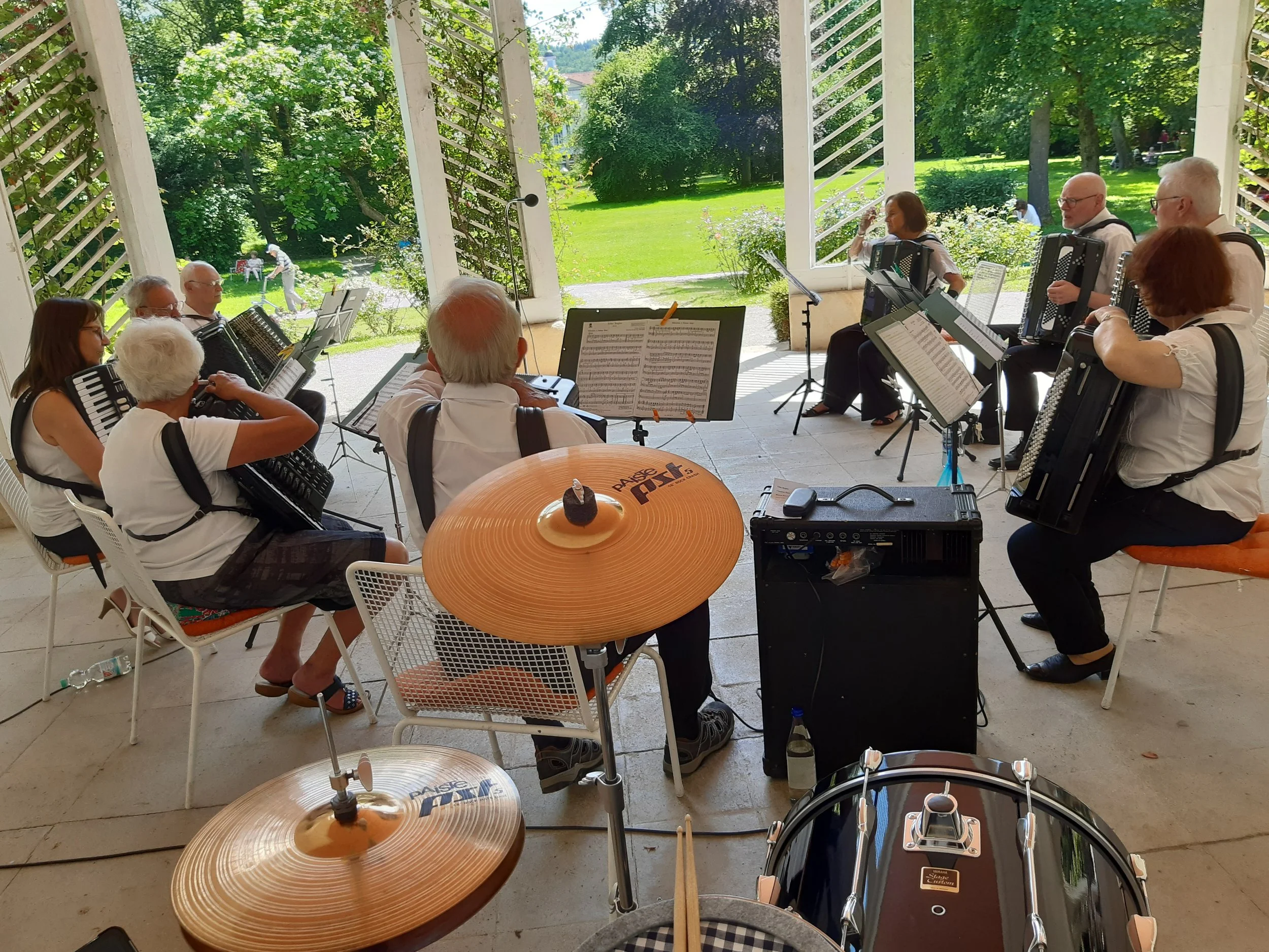 Eine Gruppe Menschen spielt ein Musikinstrumente im Freien. Sie sitzen in einem Raum mit offenen Fenstern, die einen Blick auf einen grünen Garten mit Bäumen und Wiese bieten.