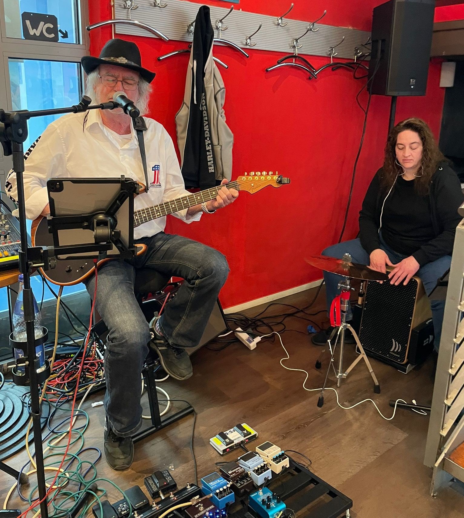 Ein älter Mann mit Brille und Hut spielt Gitarre und singt in einem Raum, während eine jüngere Frau auf einem Cajón sitzt und Musik macht. Im Vordergrund sind mehrere Effektpedale auf einem Pedalboard zu sehen. Der Raum hat eine rote Wand und eine Garderobe im Hintergrund.