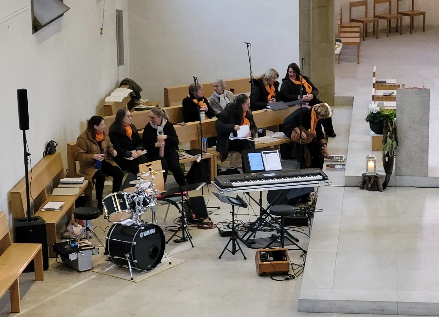 Gruppe von Frauen mit orange Scarf sitzen im Kirchenraum, einige bereiten sich auf eine musikalische Darbietung vor, mit Musikinstrumenten wie Schlagzeug, Keyboard und Mikrofonen.