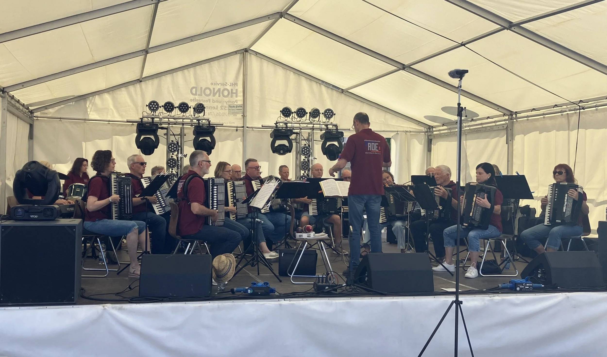 Eine Gruppe von Musikern mit Akkordeons sitzt in einem Zelt bei einem Live-Konzert, während ein Dirigent vor ihnen steht.