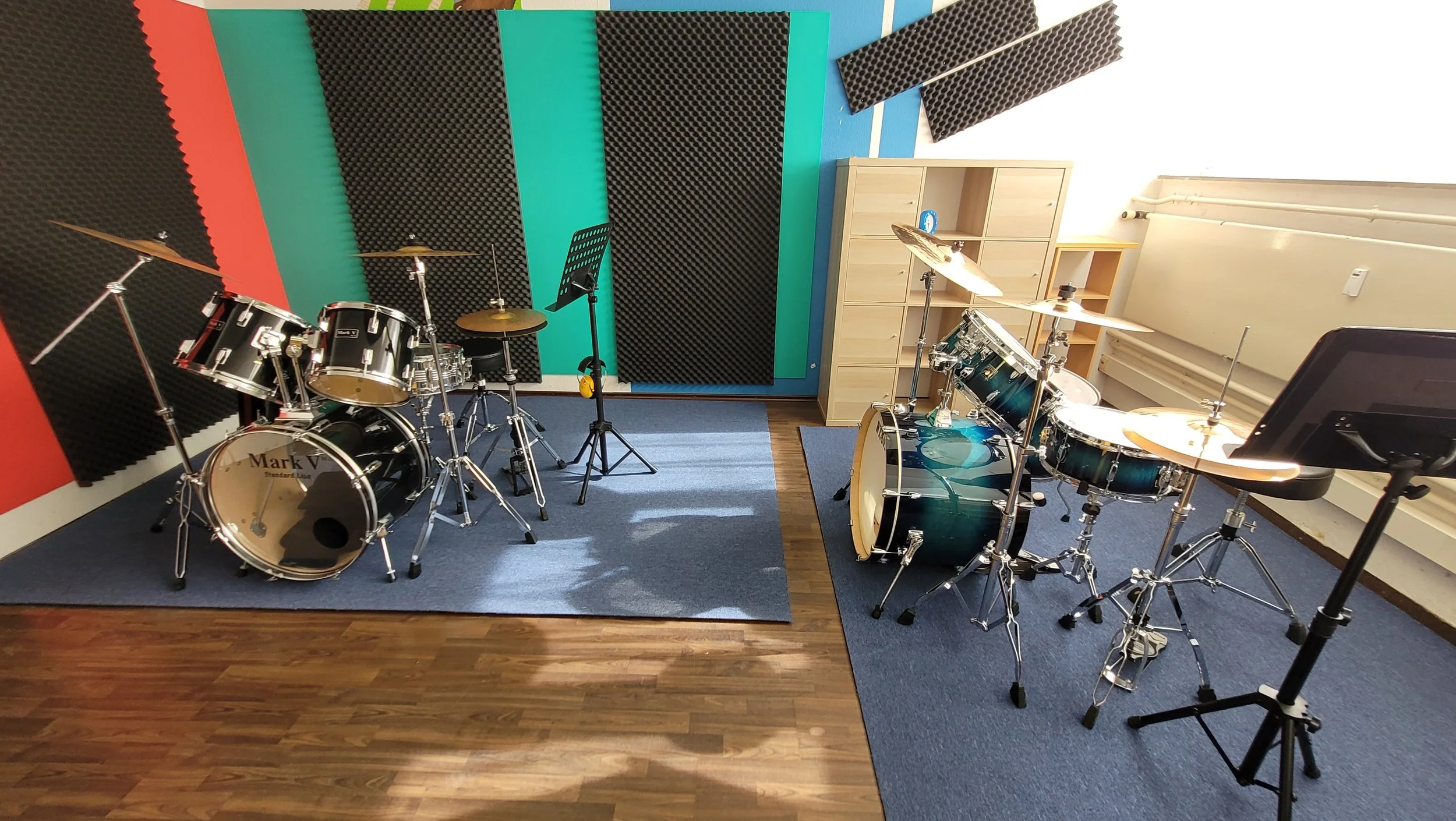 Musikzimmer mit Schlagzeug, Mikrofon, Notenständer, akustischen Wandabsorbern und einzelnen Musikinstrumenten. Die Wand ist gefliest mit akustischen Wandpaneelen, der Boden ist aus Holz mit Teppichmatten.