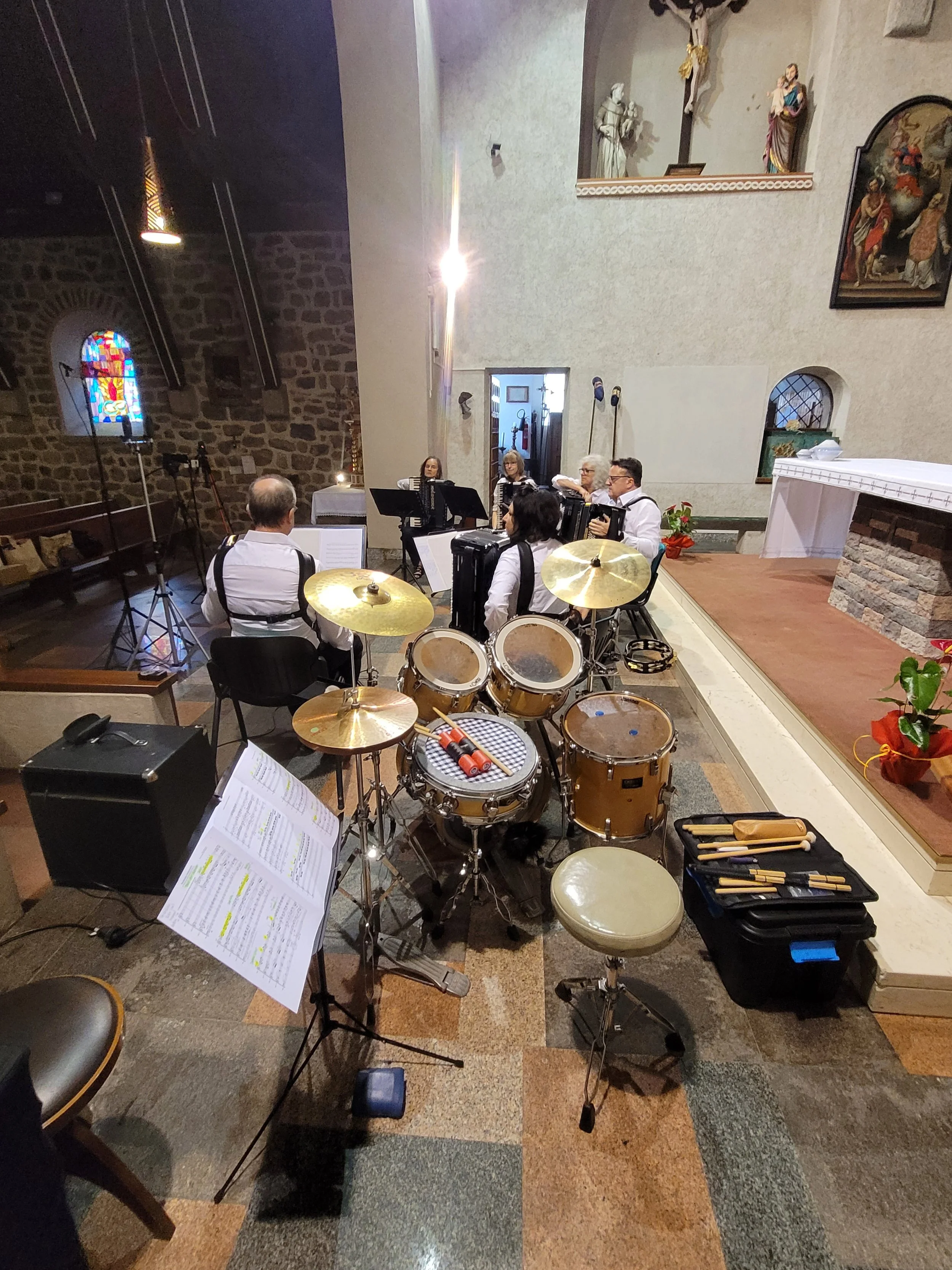Musikergruppe mit fünf Personen in einer Kirche, die Instrumente spielen, darunter eine Trommel und Akkordeons, im Hintergrund religiöse Kunstwerke und Statuen.