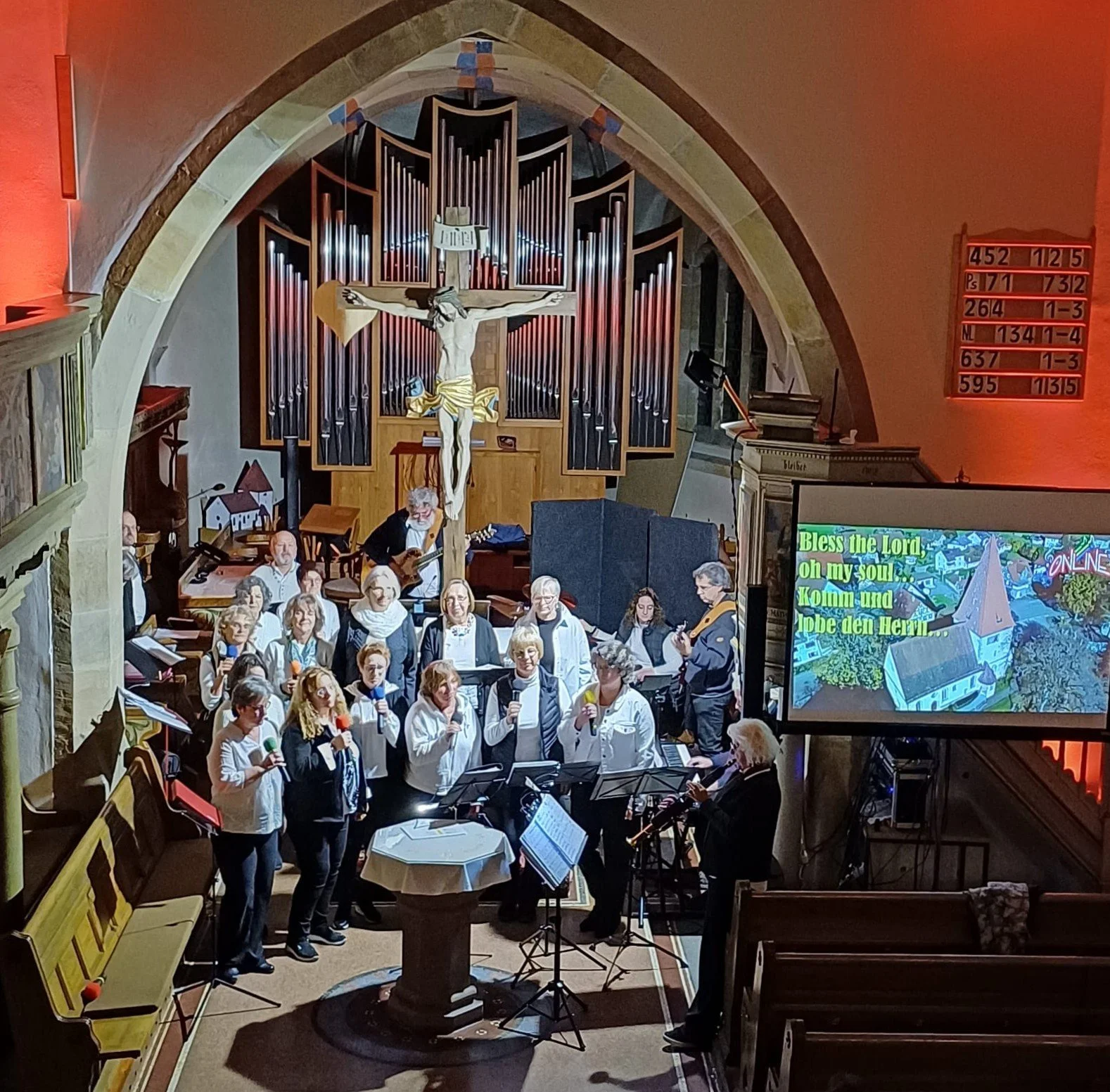 Innenaufnahme einer Kirche mit einem Kreuz und einer Orgel im Hintergrund. Eine Gruppe von Menschen singt mit Mikrofonen, begleitet von Musikern. Es gibt einen Bildschirm mit einem deutschen Liedtext und eine elektrisch angezeigte Nummerntafel.