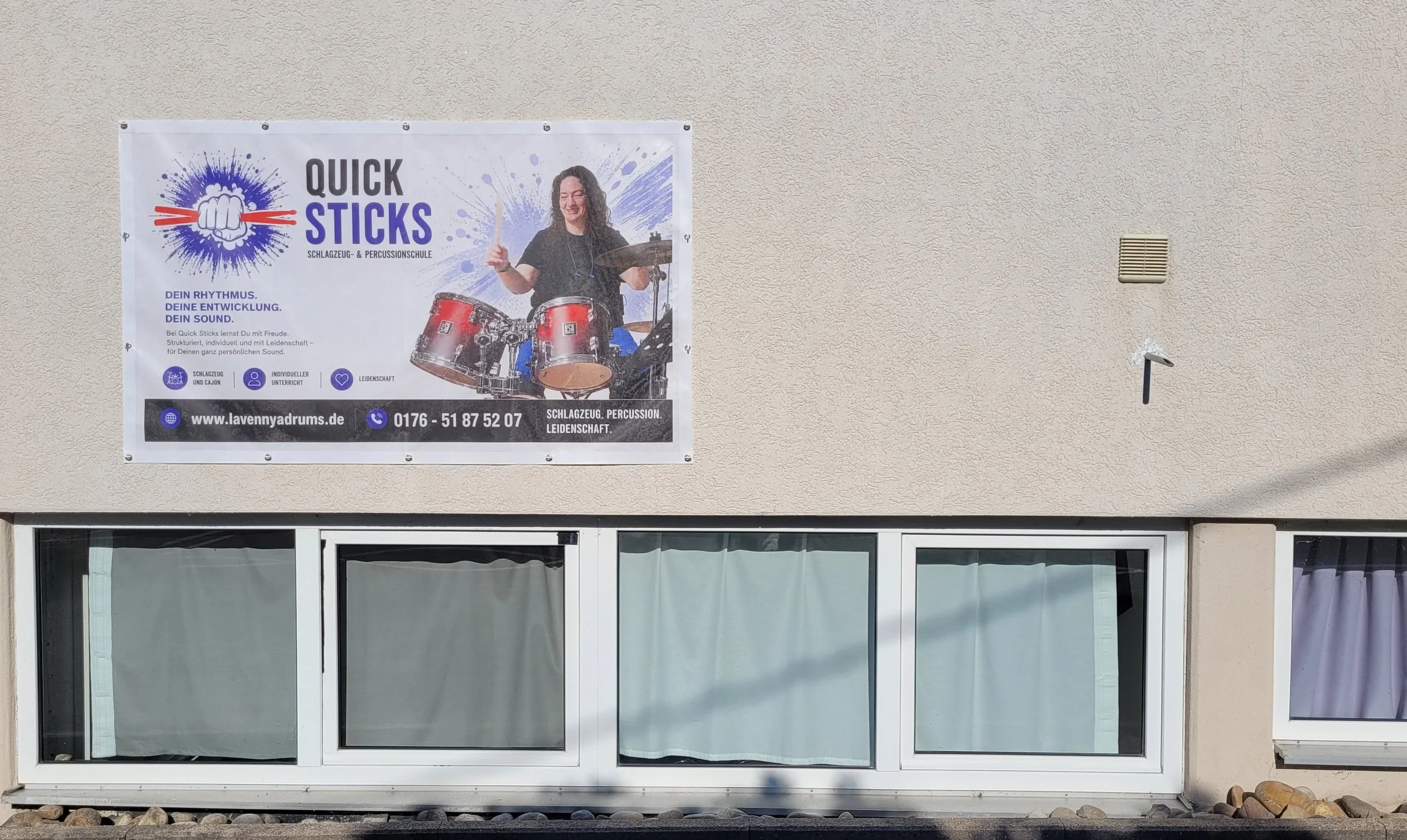 Werbebanner für eine Trommel- und Percussionschule mit einem lachenden Mädchen, das auf einer roten Trommel spielt, neben einem Fenster in einem weißen Gebäude.