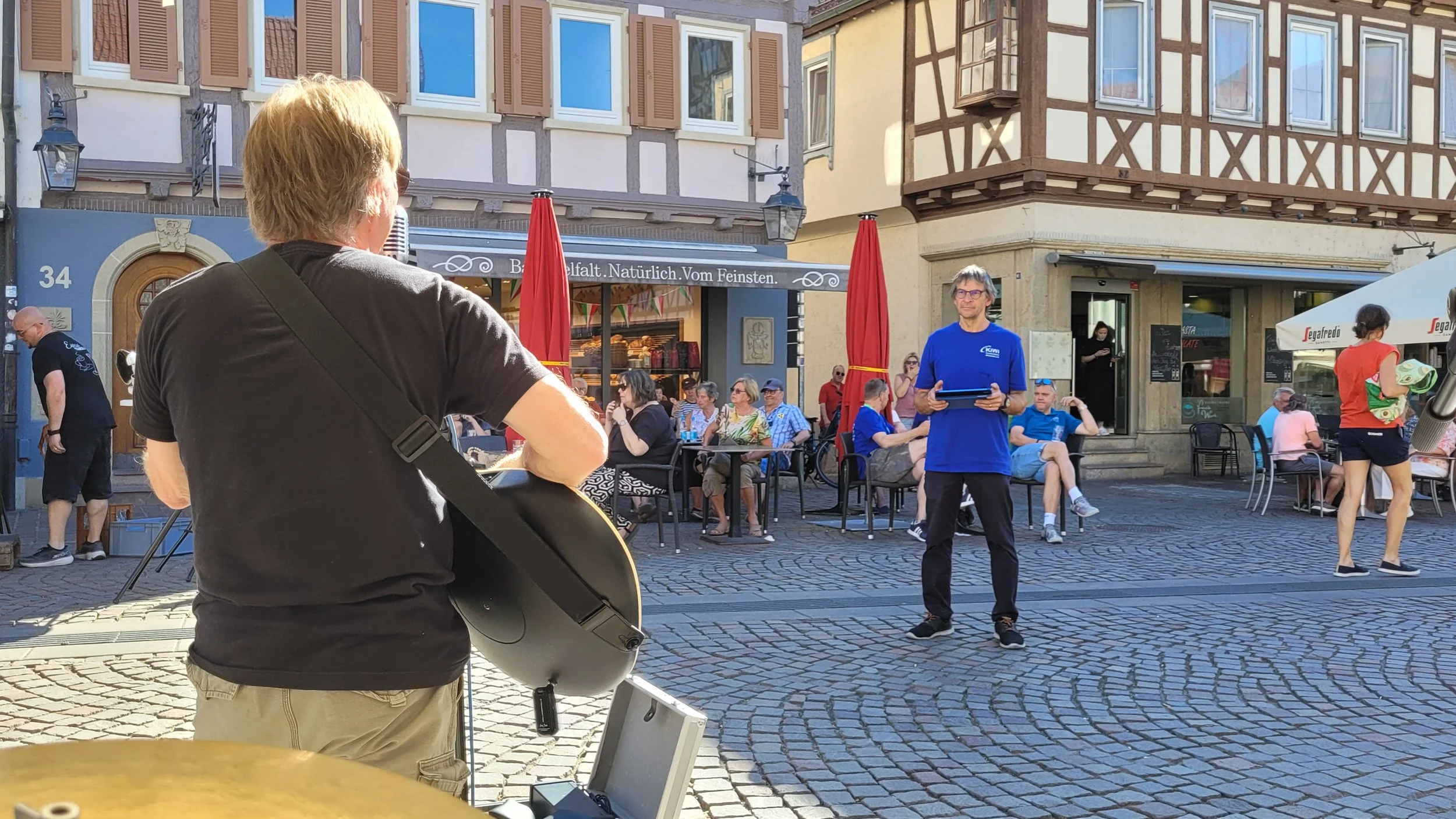 Menschen sitzen und stehen auf einem belebten Stadtplatz, einige mit Getränken, und einer spielt Gitarre. Es gibt Cafés und historische Gebäude im Hintergrund.