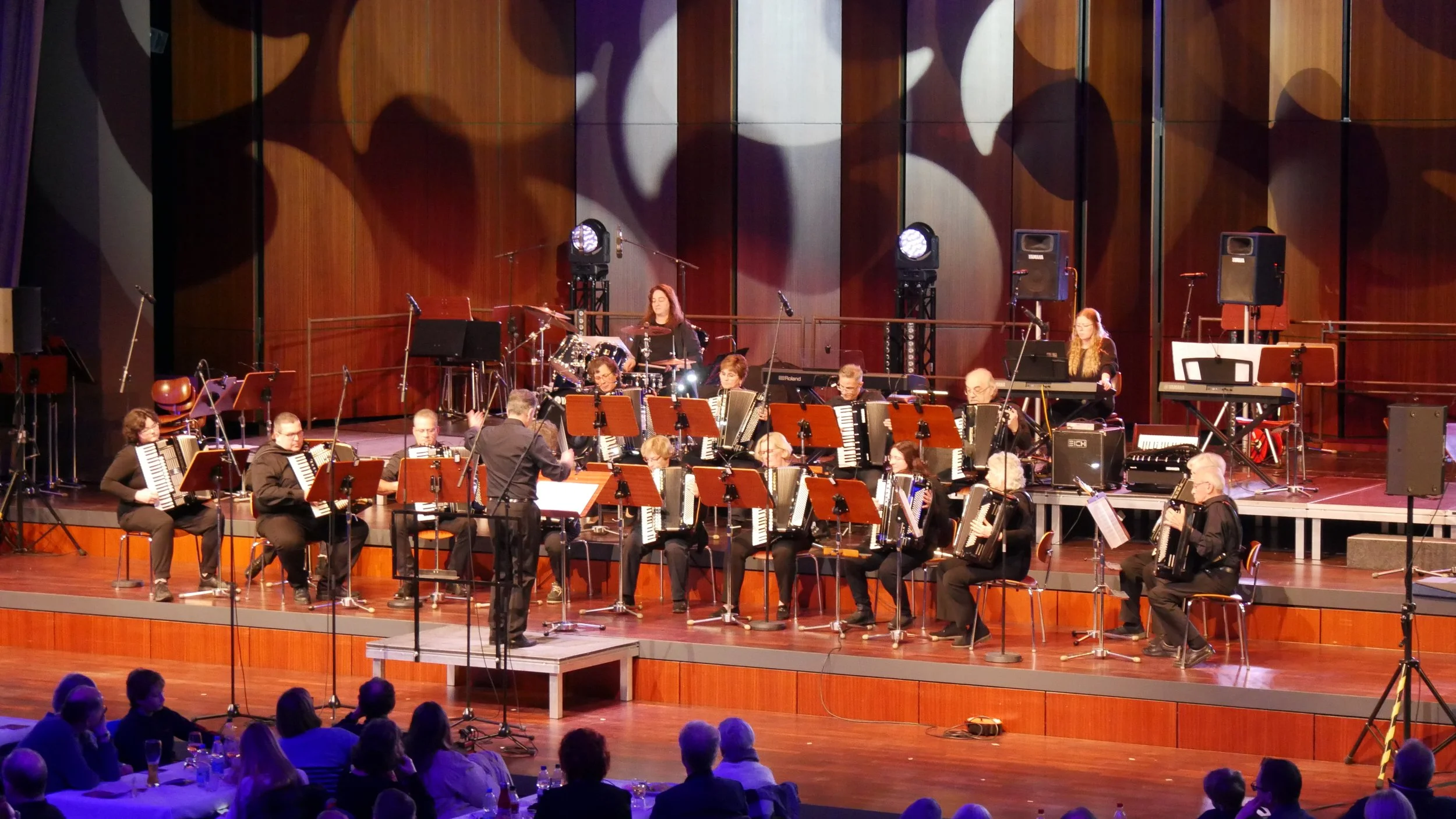 Eine große Orchestergruppe spielt auf einer Bühne in einem Konzertsaal. Das Orchester besteht aus Musikern, die verschieden Instrumente wie Akkordeon, Schlagzeug, Klavier und andere spielen. Das Publikum sitzt im unteren Bereich und beobachtet die Aufführung. Die Bühne ist mit Scheinwerfern beleuchtet, und die Wände sind mit Holzpaneelen verkleidet.