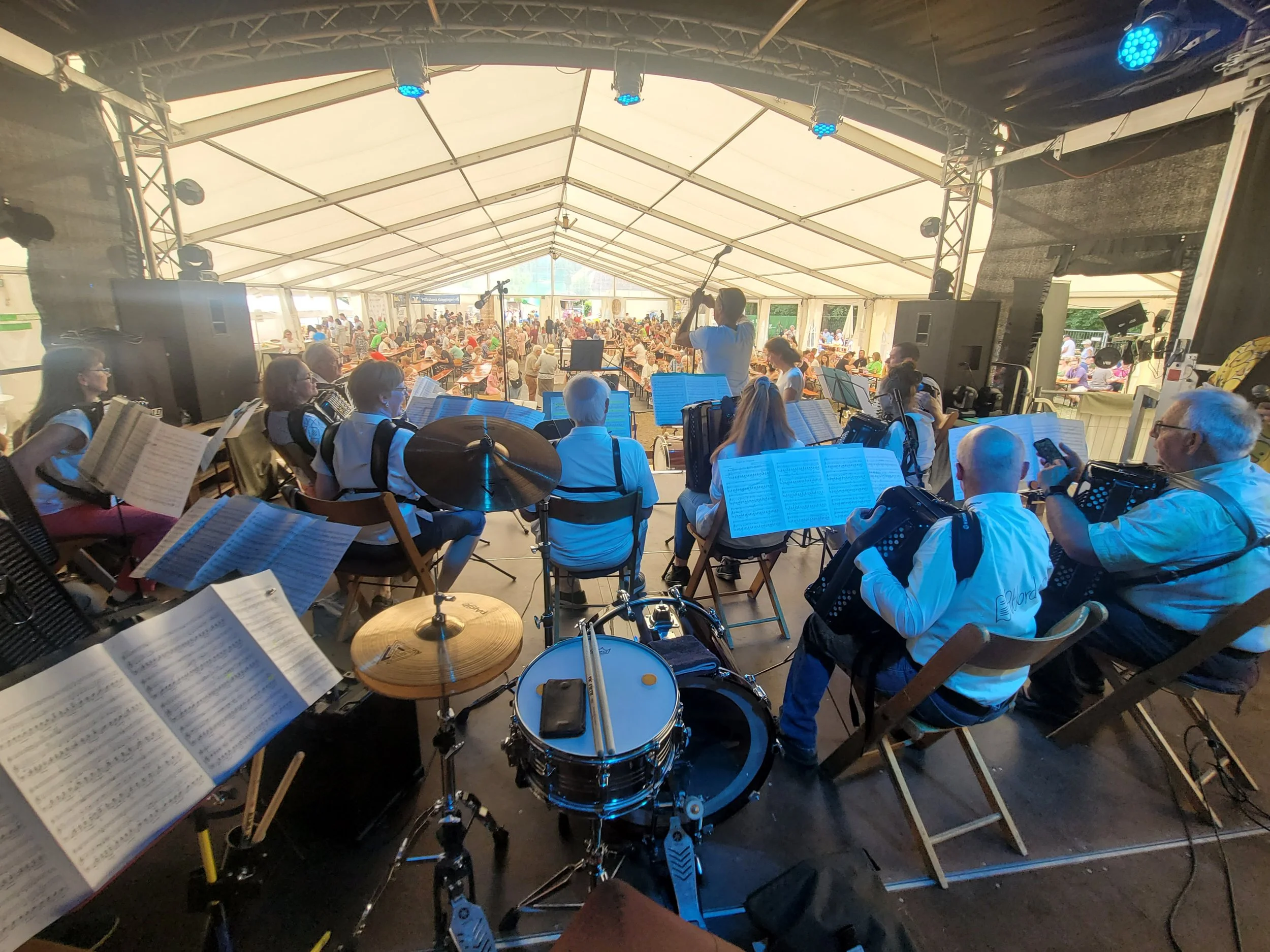 Ein großes Zelt, in dem eine Orchester- oder Musikgruppe mit Musikern auf der Bühne spielt, während das Publikum in den Sitzgelegenheiten im Inneren des Zeltes sitzt und das Konzert genießt.