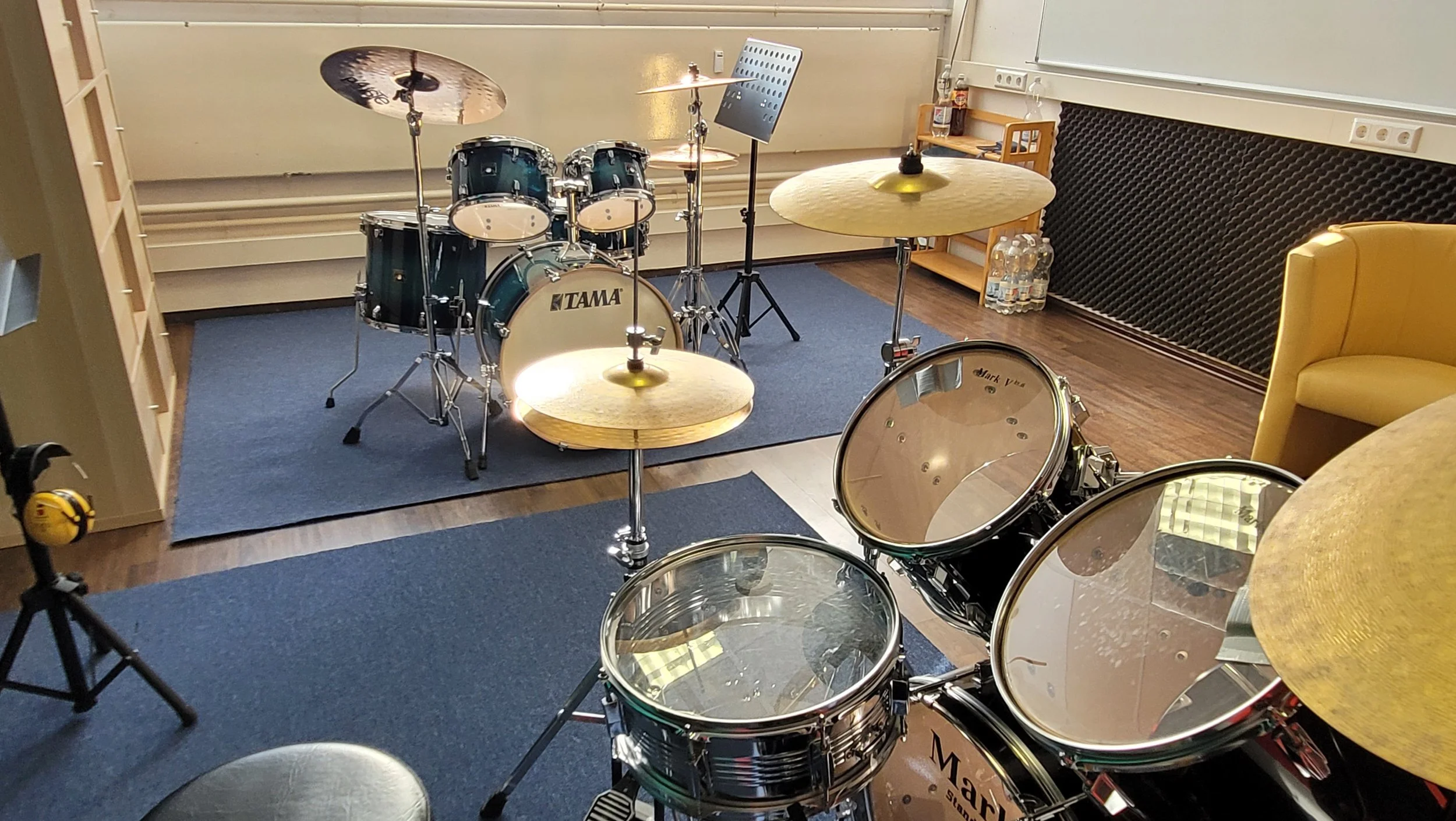 Musikstudio mit Schlagzeug, Cymbals und Sitzgelegenheiten auf einem blauen Teppichboden in einem Raum.