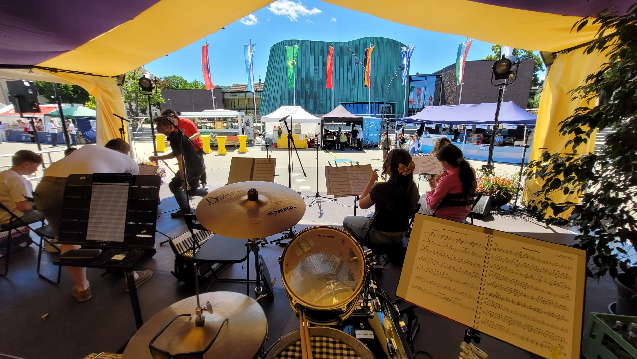 Blick aus einer Bühne auf einen offenen Platz, im Hintergrund bunter Flaggen, moderne Gebäude, und Besucher im Freien bei einem Festival.