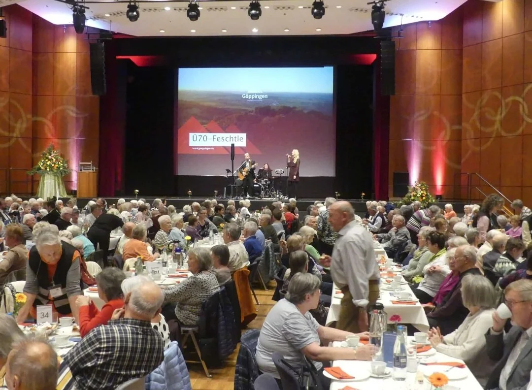 Große Veranstaltung mit Menschen, die an Tischen sitzen, eine Bühne mit Musikern und Sängern, ein großer Bildschirm im Hintergrund zeigt eine Landschaft und den Text 'Ü70-Festchle'.