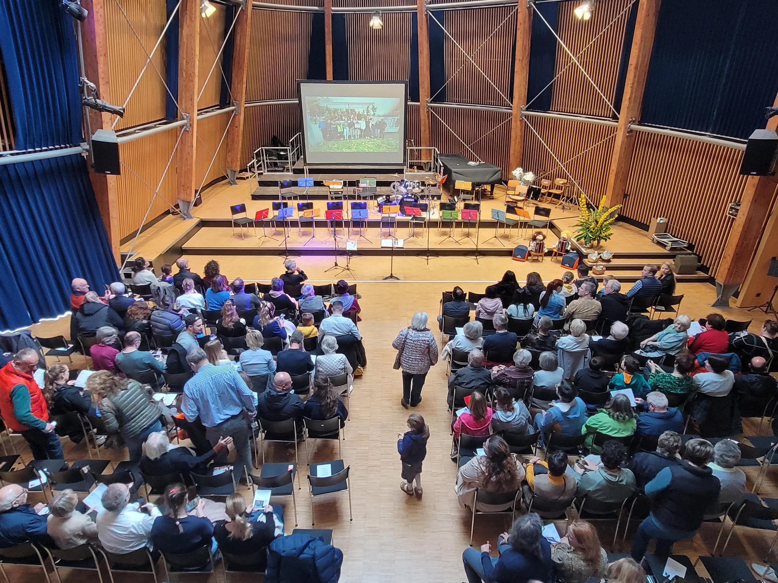 Ein Konzertsaal mit Publikum, Sitzreihen, einem großen Bildschirm, einigen Instrumenten. Das Foto zeigt den Beginn einer Veranstaltung oder ein Konzert.