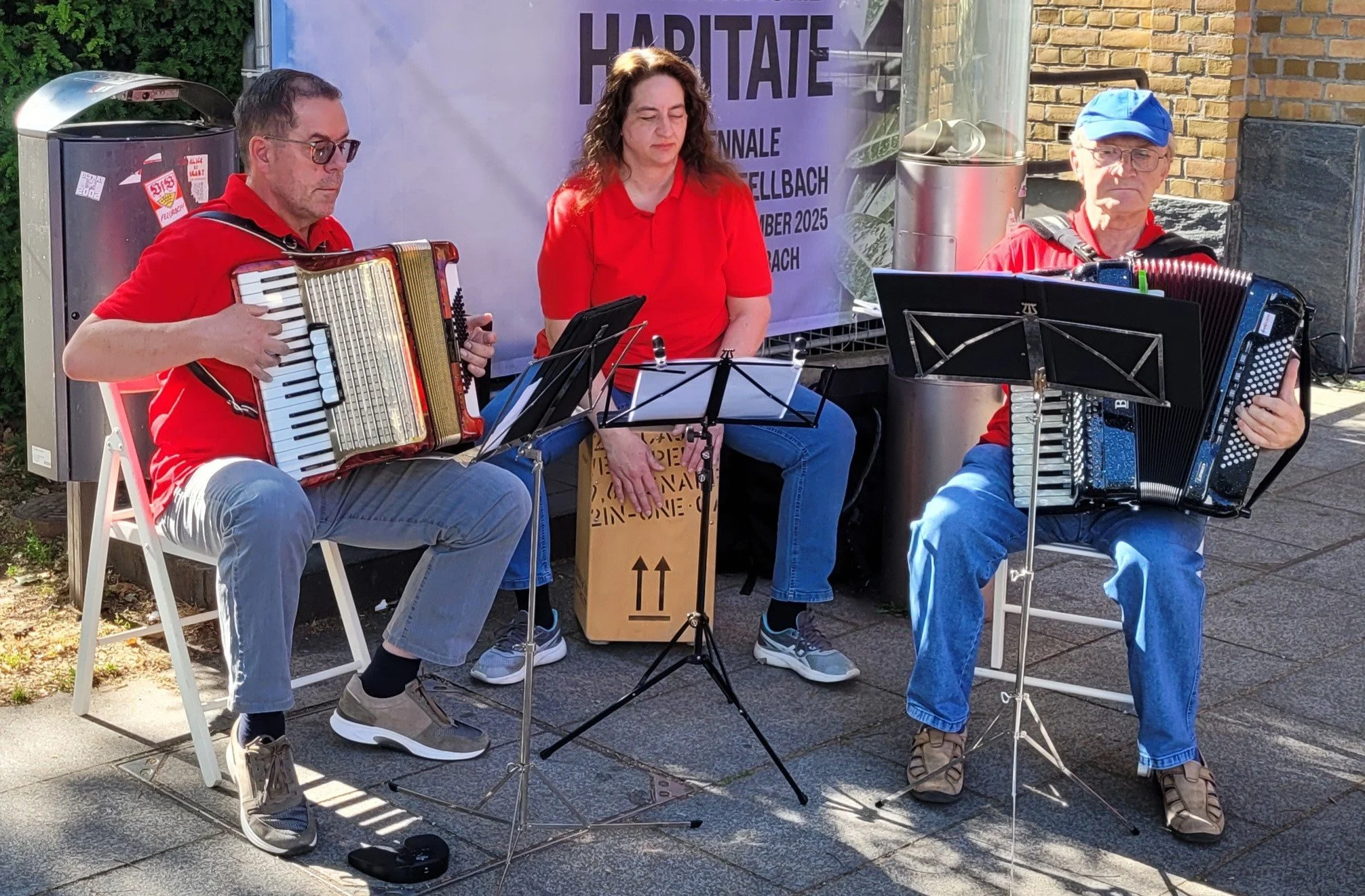 Drei Musiker, zwei Männer und eine Frau, spielen auf der Straße. Sie tragen rote Hemden. Die Männer spielen Akkordeon, die Frau sitzt in der Mitte und spielt Cajon.