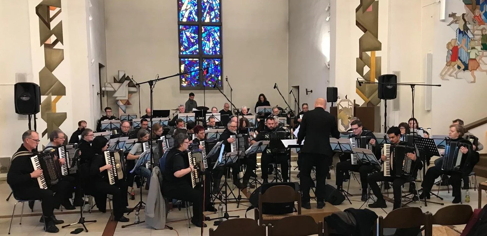 Orchester mit Männern und Frauen, die Akkordeon spielen, dirigiert von einem Mann, vor einer Kirchenfenster mit buntem Glas, in einer Kirche mit weißen Wänden und künstlerischen Wanddekorationen.