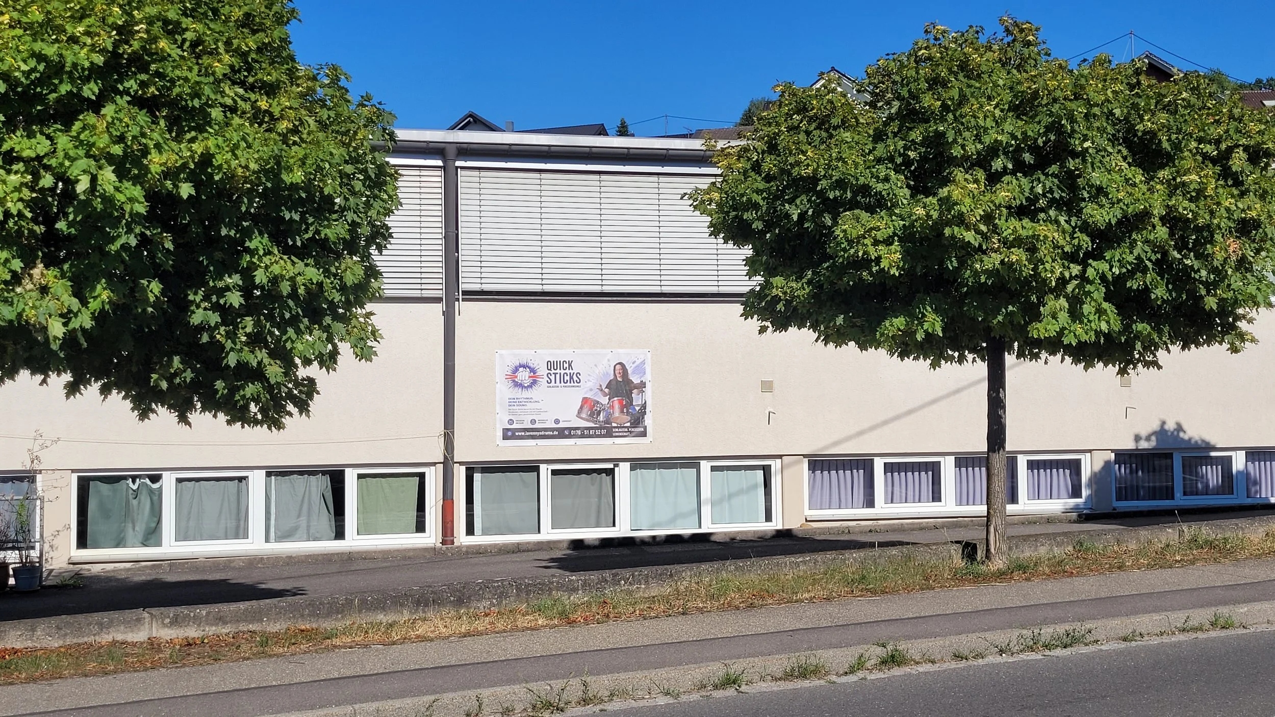 Zweistöckiges Gebäude mit Werbebanner und mehreren Fenstern, umgeben von Bäumen, bei sonnigem Wetter.