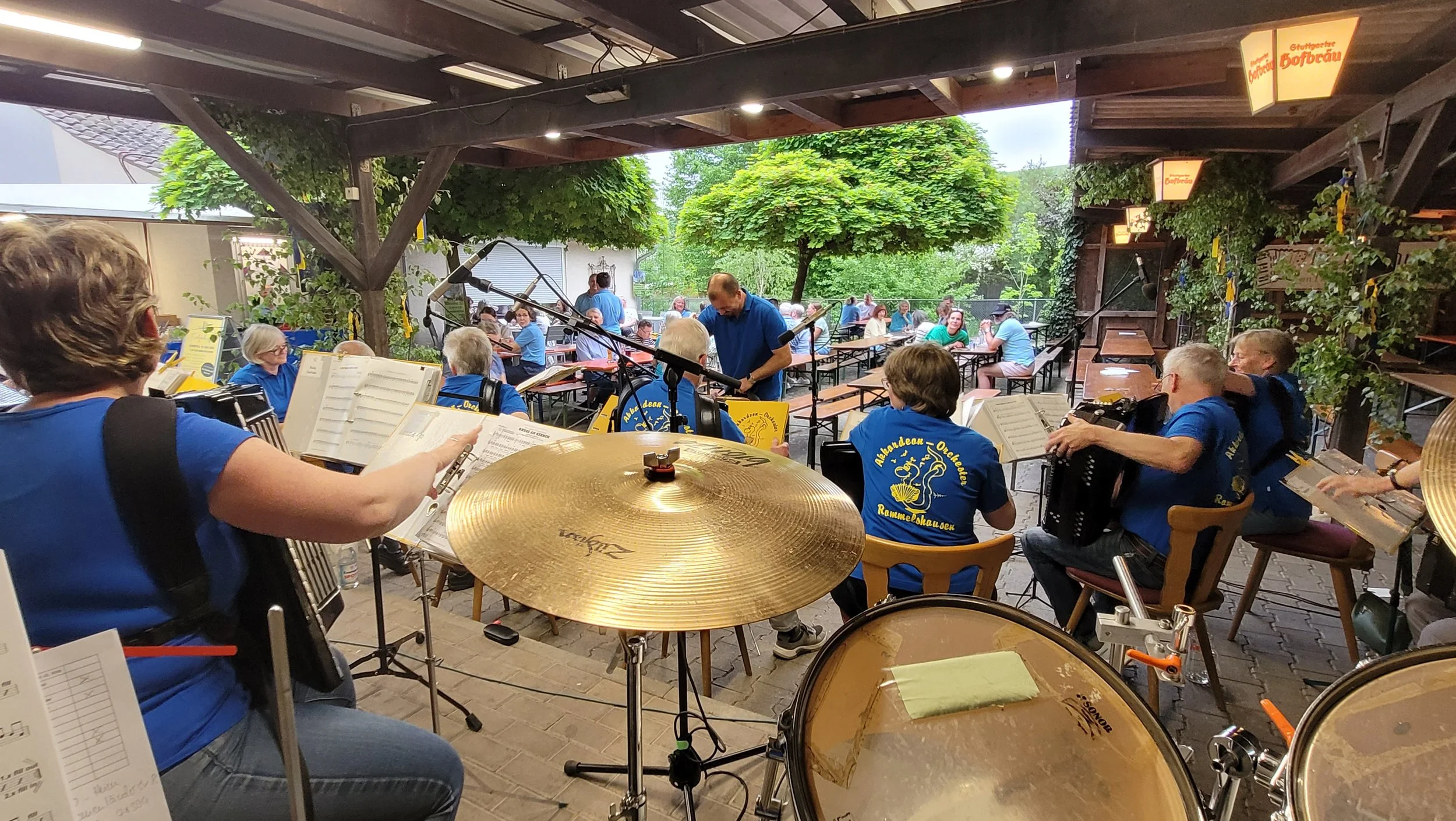 Blick auf eine Live-Musikperformance im Freien mit Musikern, die auf einer gedeckten Terrasse spielen, während draußen Menschen an Tischen sitzen und die Musik genießen, umgeben von grünen Bäumen.