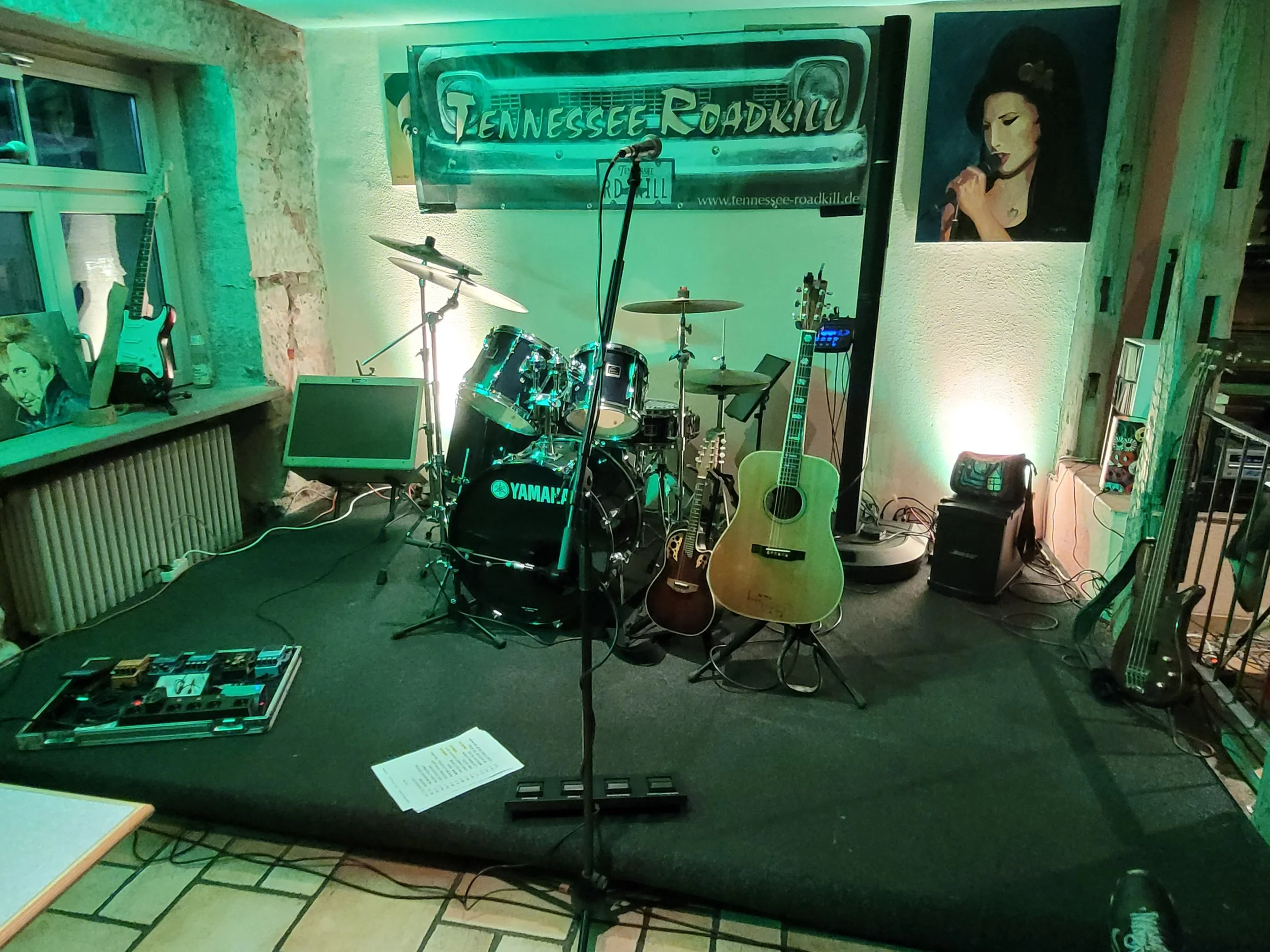 Musik-Bühne mit Drums, Gitarren und Mikrofon, dekoriert mit Tennessee Roadkill Banner, auf einer kleinen Bühne, umgeben von Instrumenten und musikalischer Ausrüstung in einem gemütlichen Raum.