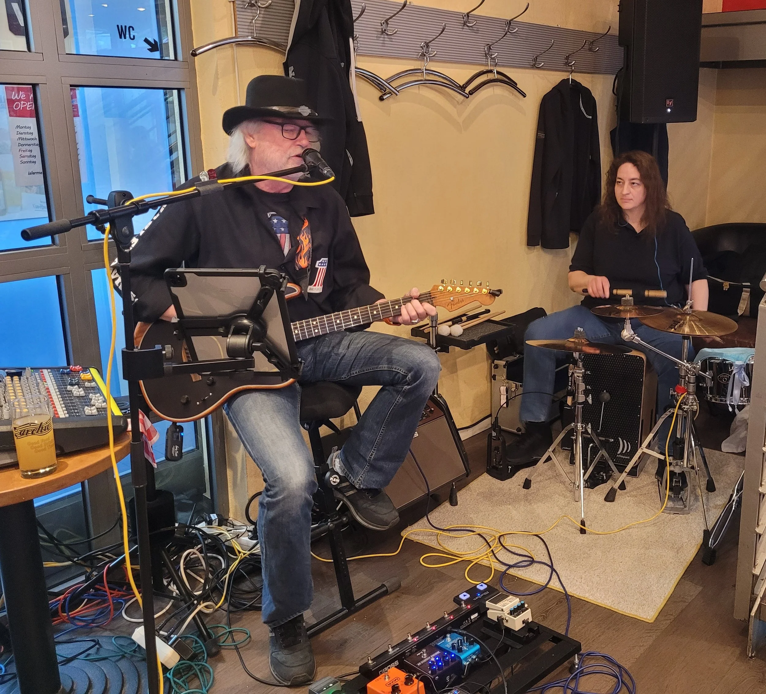 Auf ener kleinen Bühne sitzt Mann und spielt Gitarre und singt. Neben ihm sitzt eine Frau mit lockigen Haaren , die auf einem Schlagzeug spielt.