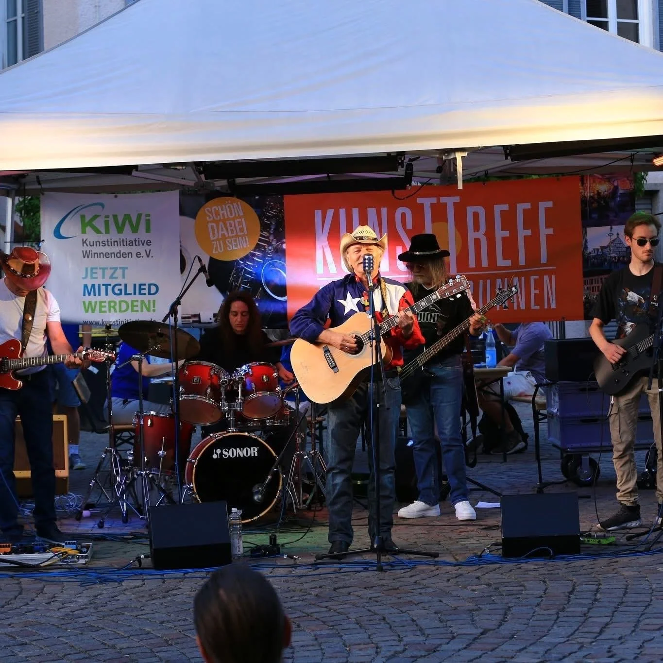 Eine Straßenmusikgruppe spielt auf einer Bühne unter einem weißen Pavillon. Der Sänger trägt einen Cowboyhut und Gitarre, während andere Musiker Gitarre und Schlagzeug spielen. Im Hintergrund sind Banner mit Aufdrucken in Deutsch, einer Werbung für eine Kunstrichtung und eine Veranstaltung.