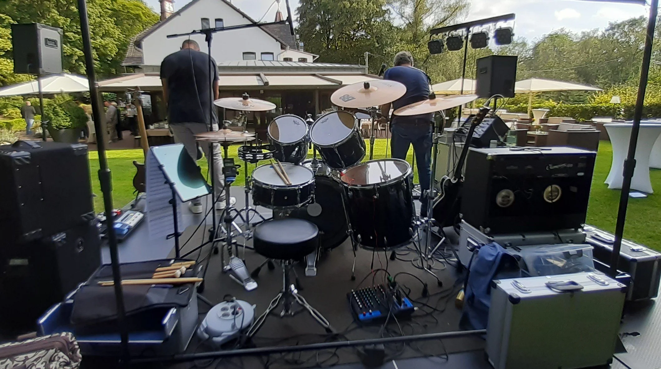 Auf einer Bühne im Freien stehen zwei Musiker, die auf eine Aufführung vorbereitet sind. Es gibt ein Schlagzeug, Verstärker und andere Musikausgerätschaften. Im Hintergrund sind Bäume und ein Haus zu sehen.