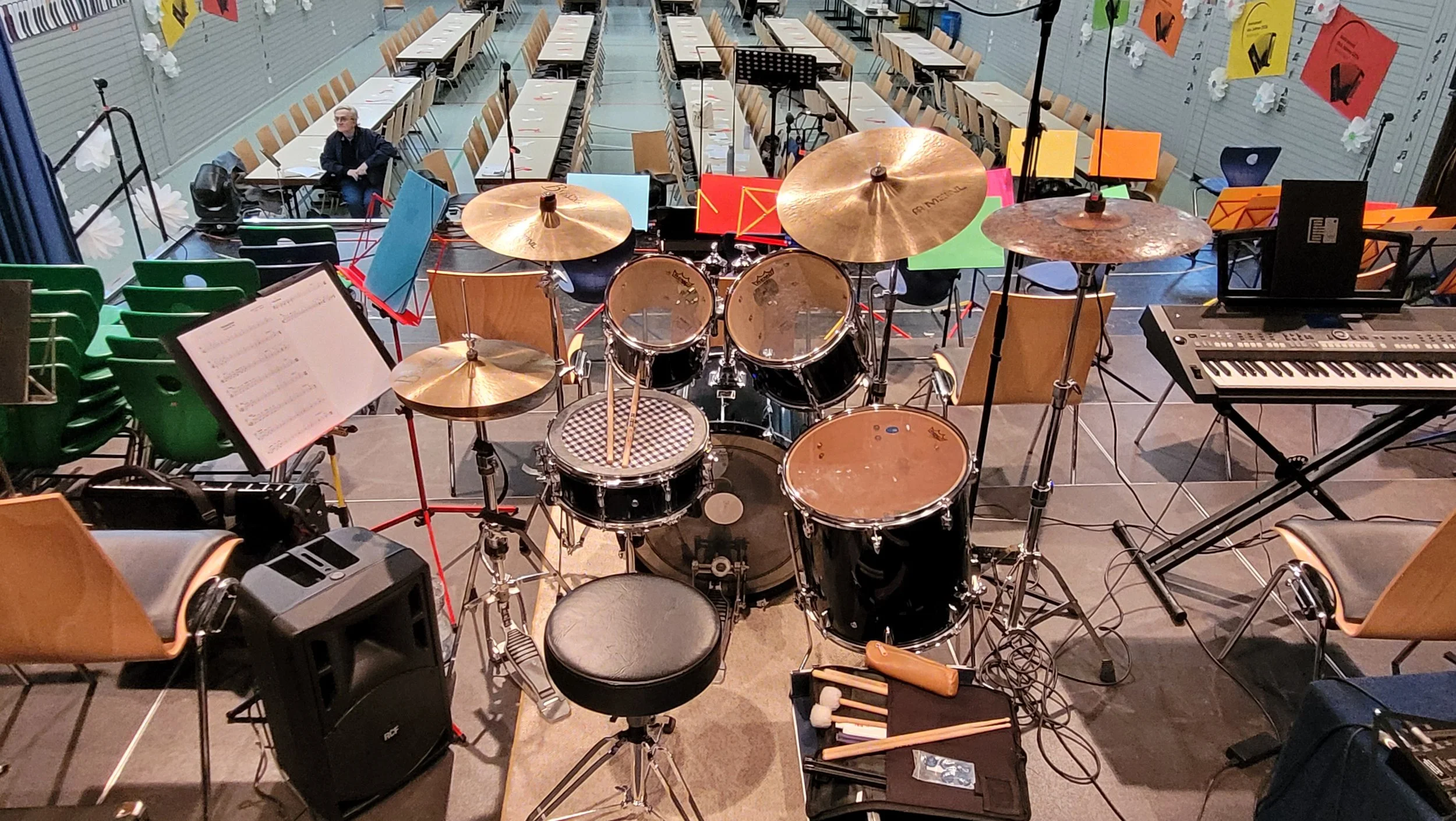 Blick von der Bühne auf ein leeres Klassenzimmer mit Tischen, Stühlen, Musikinstrumenten und einem Sitzhaltunggericht im Vordergrund.