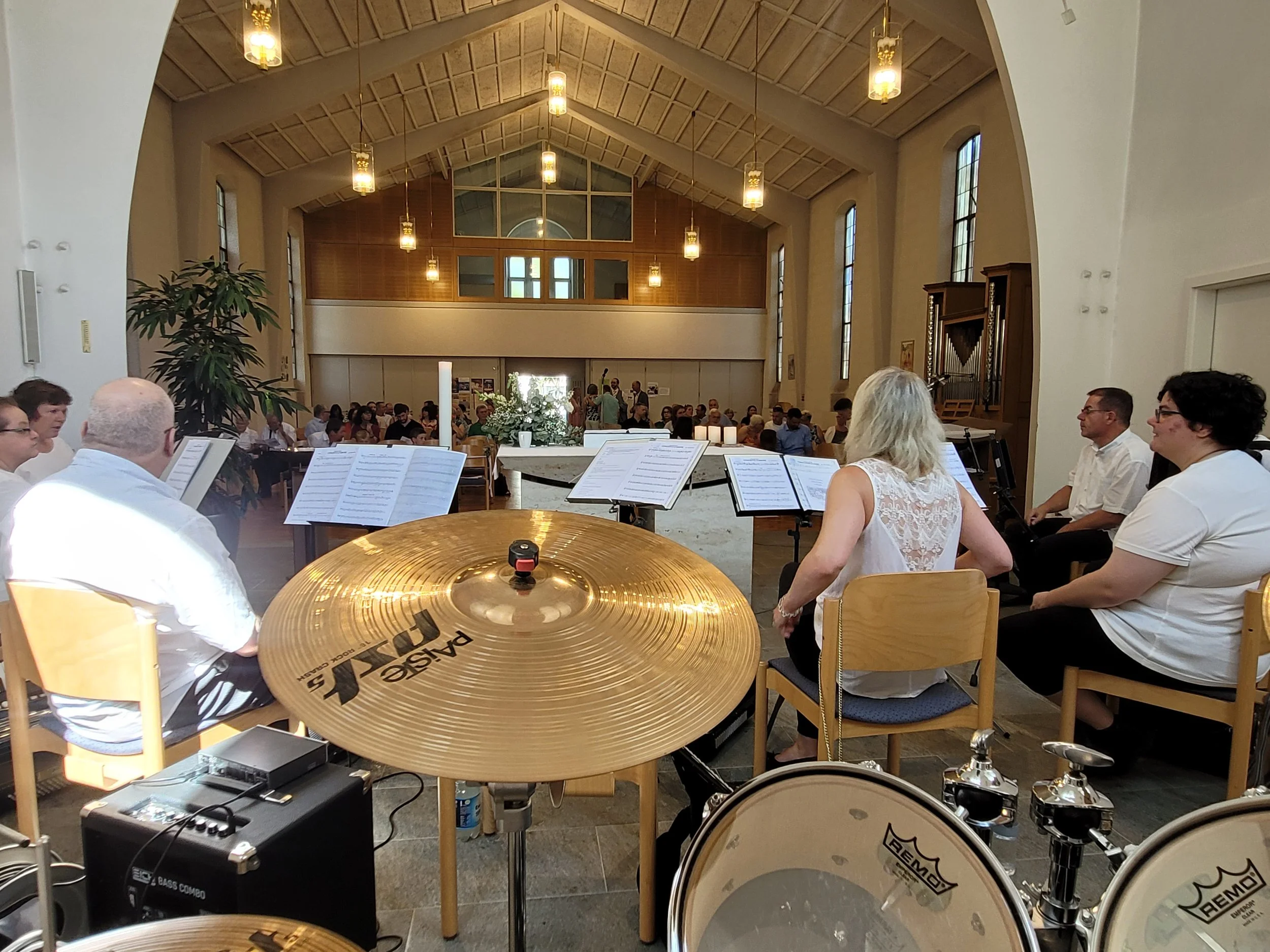 Innenaufnahme eines Kirchenraums während eines Konzerts. Im Vordergrund sind Schlagzeug und Notenständer sichtbar, im Hintergrund ein Chor oder Orchester, das in einer Kirche mit hohen Fenstern und Holzdecke auftritt.