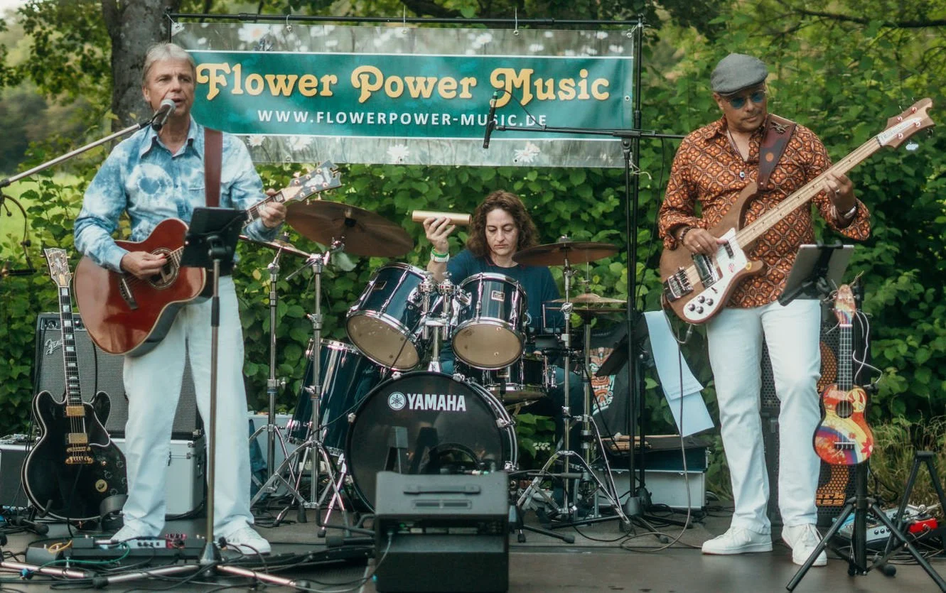 Drei Musiker auf einer Bühne im Freien, einer mit Gitarre singend, einer am Schlagzeug und einer am Bass, mit einem Banner im Hintergrund, das "Flower Power Music" und eine Website zeigt.