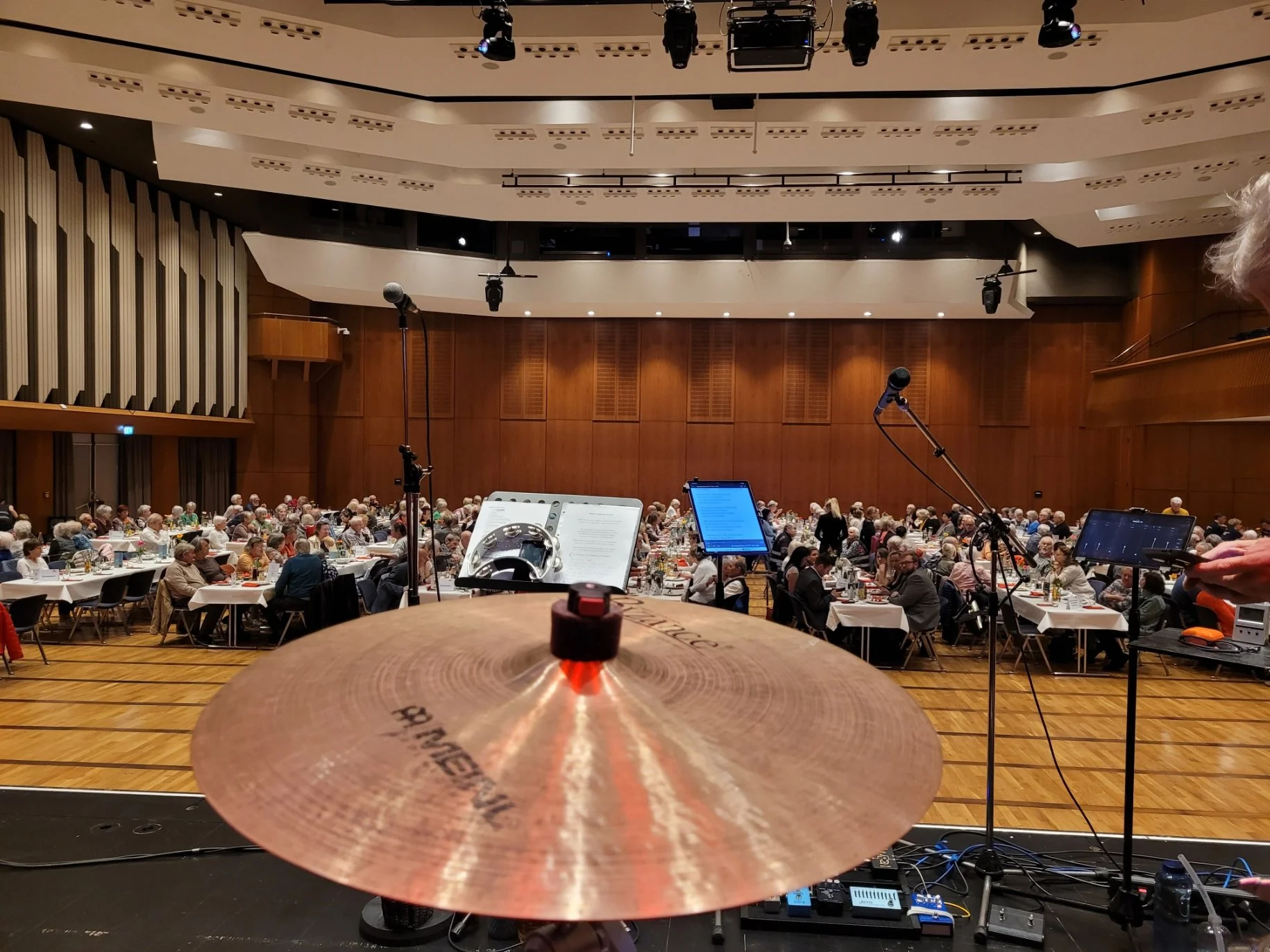 Blick von einem Schlagzeug auf eine große Konferenz- oder Konzertveranstaltung mit vielen Sitzplätzen und Menschen im Saal. Im Hintergrund sind die Bühne und die technische Ausstattung sichtbar.