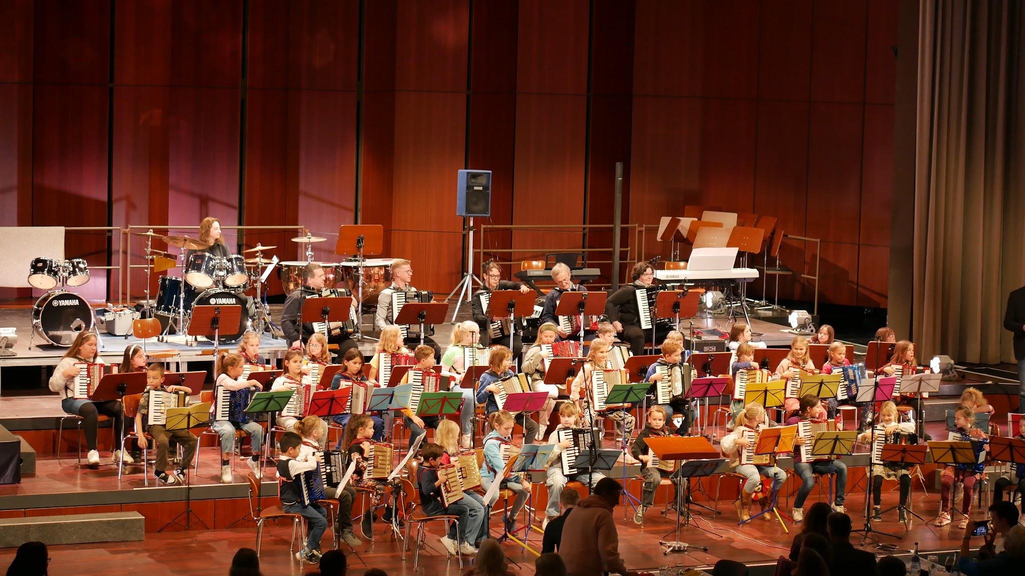 Ein Kinderorchester auf einer Bühne mit vielen Kindern, die Musikinstrumente spielen, begleitet von einem Erwachsenen, im Hintergrund stehen Musikinstrumente wie Schlagzeug, Keyboard und Klaviere, vor einer roten Holzwand.