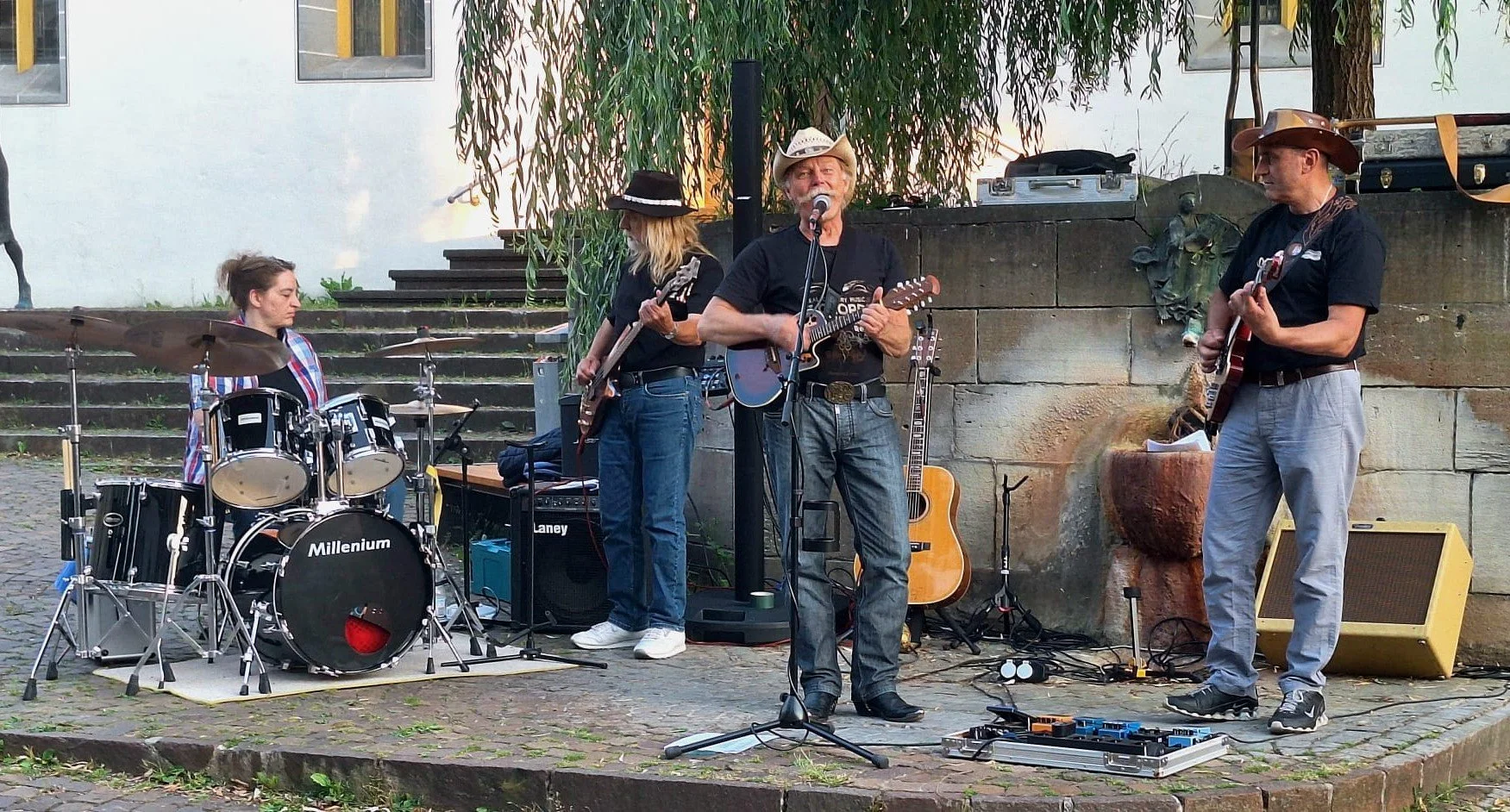 Vier Musiker spielen auf einer Bühne im Freien. Es gibt eine Schlagzeugerin, eine Sängerin, und zwei Gitarristen. Sie tragen Cowboy-Hüte und sind umgeben von Musik-Equipment.