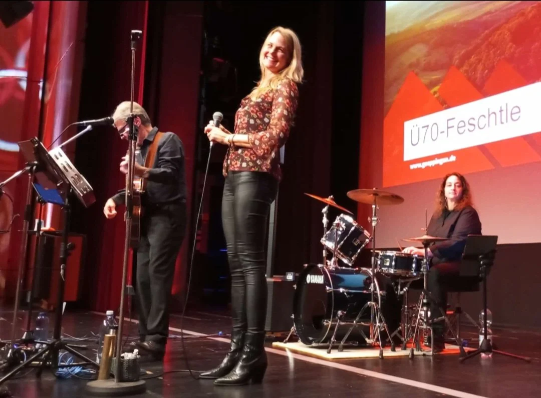 Musiker auf einer Bühne während einer Veranstaltung, eine Frau singt in ein Mikrofon, ein Mann spielt Gitarre, eine Frau sitzt am Schlagzeug, im Hintergrund eine große Leinwand mit dem Text 'Ü70-Festtle'.