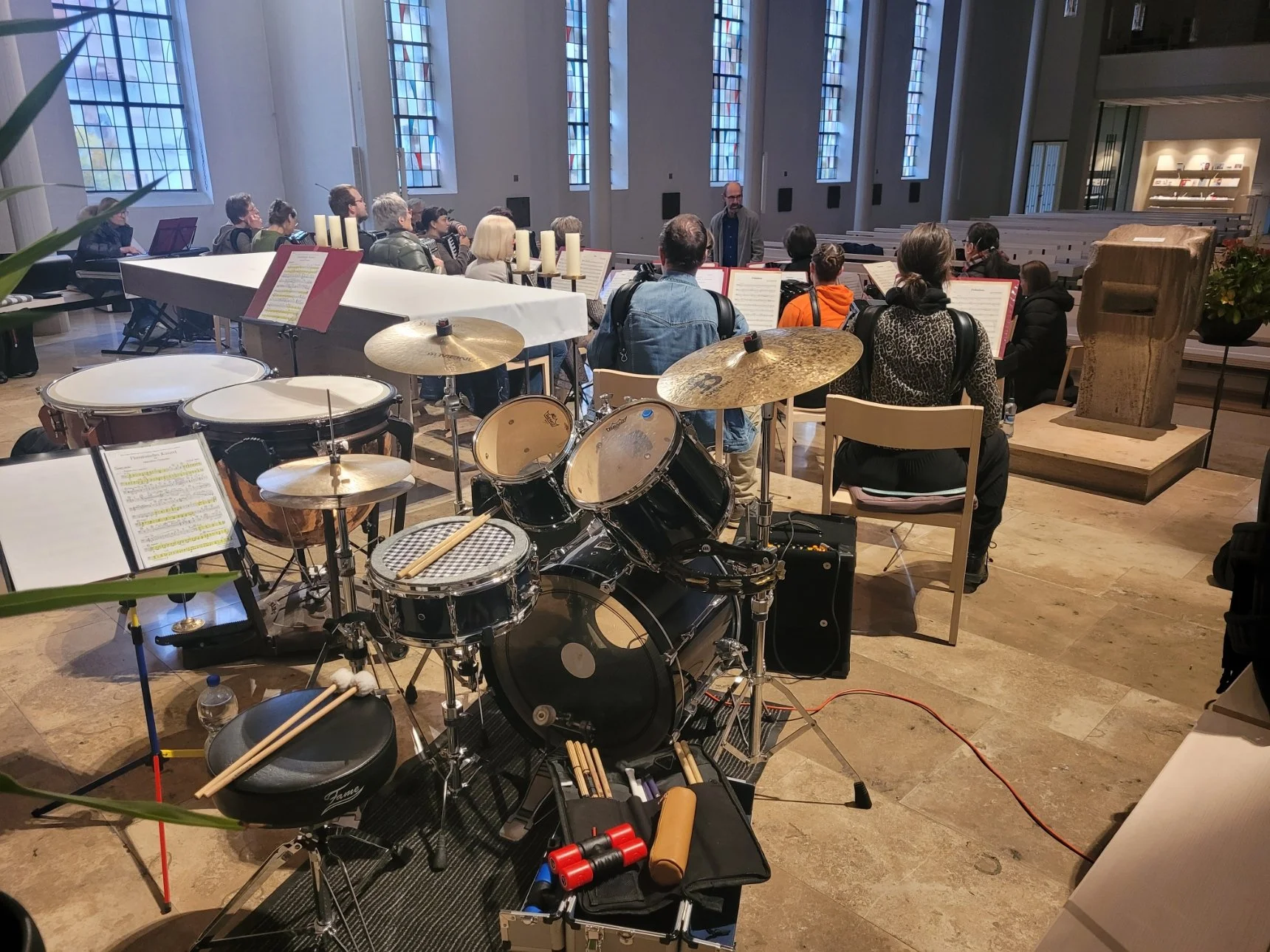 Innenaufnahme einer Kirche oder Konzerthalle, in der eine Gruppe von Menschen an einem Chorprojekt teilnimmt, mit einem Dirigenten. Im Vordergrund steht ein Schlagzeug, im Hintergrund sind Bänke und bunte Kirchenfenster sichtbar.
