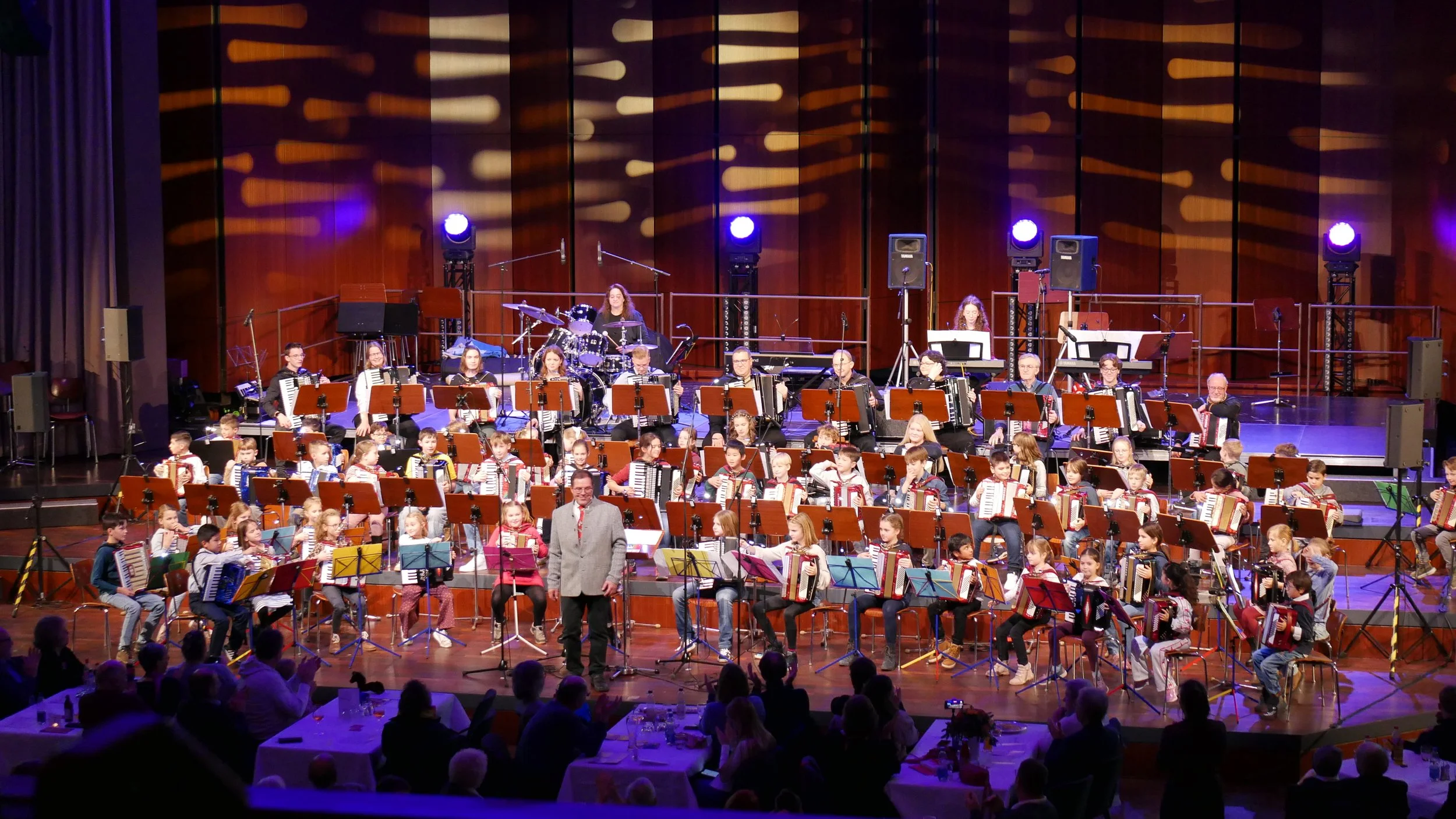 Ein Orchester mit jungen Musikern und Erwachsenen auf einer Bühne während eines Auftritts, mit vielen Sitzplätzen und buntem Licht.